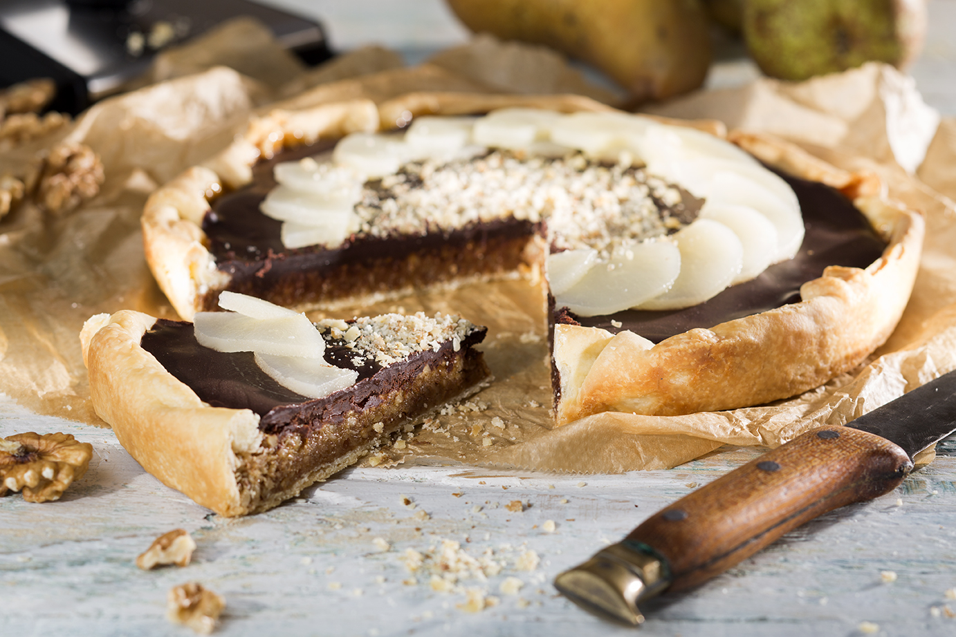 Salzkaramell-Walnusstarte mit Birnen Eine Schokoladen-Birnen-Tarte mit gehackten Nüssen auf Backpapier, ein Stück wurde herausgeschnitten, neben einem Messer