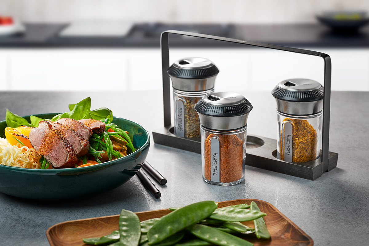 Ein stilvolles Gewürzstreuer-Set mit verschiedenen Gewürzen, platziert neben einer Schüssel mit asiatischer Küche, inklusive Fleisch und Gemüse auf einem Tisch.