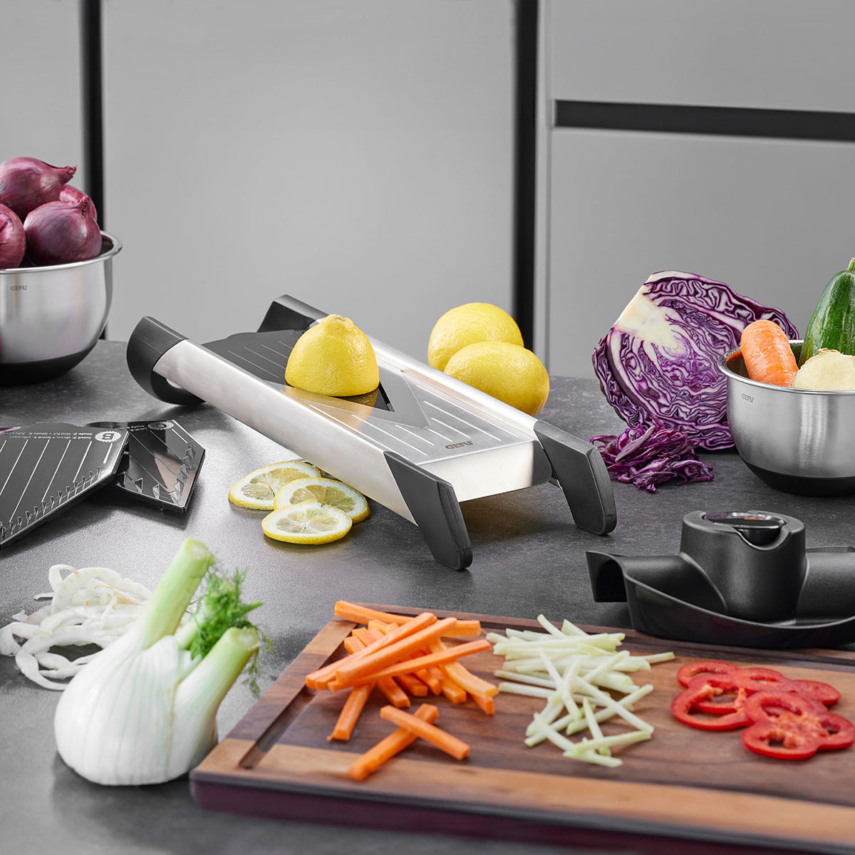 Küchenreibe mit frischem Gemüse und Obst auf Arbeitsplatte Eine Küchenreibe umgeben von geschnittenem Gemüse und Obst auf einer grauen Arbeitsplatte. Im Hintergrund befinden sich Zwiebeln, Zitronen und ein Schneidebrett mit verschiedenen Gemüsesorten.