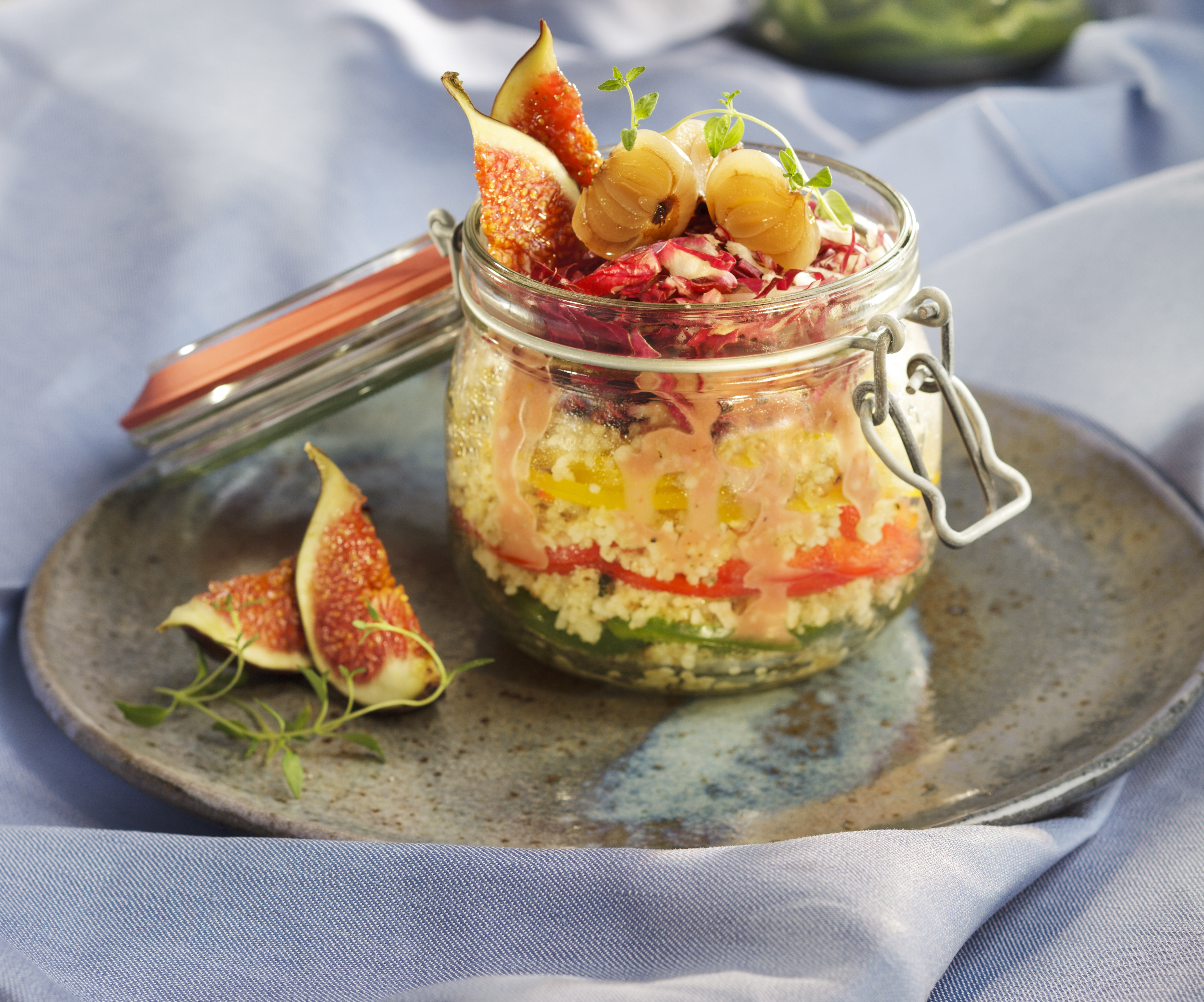 Couscous Paprika Schichtsalat mit Feigen-Vinaigrette Ein dekoratives Einmachglas mit Couscous-Salat, garniert mit frischen Feigen und einem cremigen Dressing, auf einem blauen Tuch platziert.