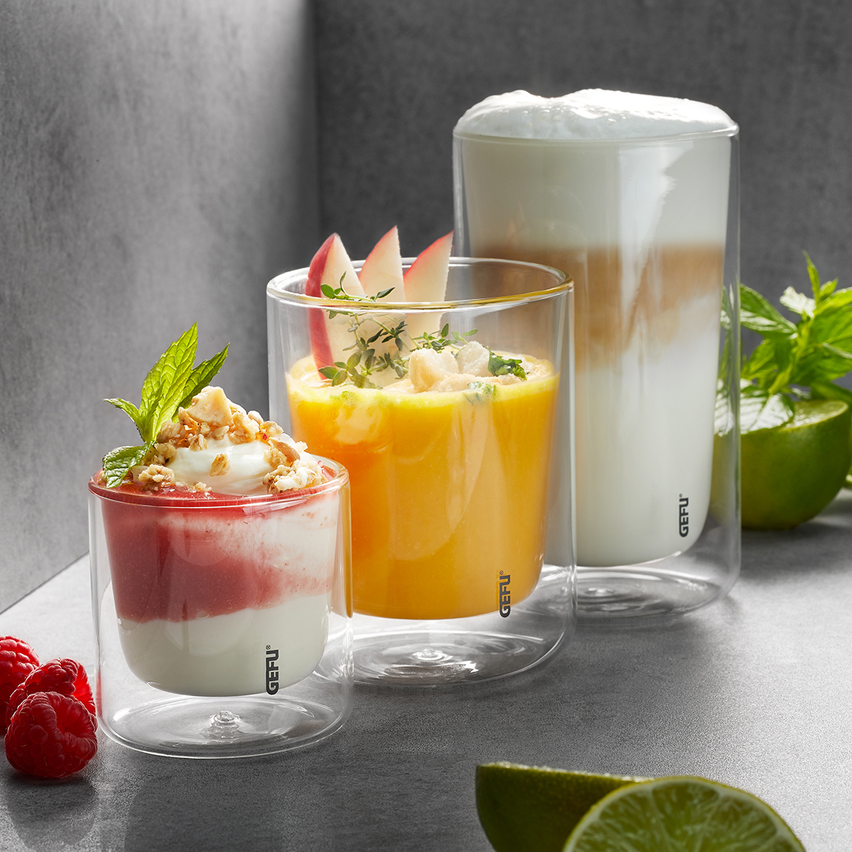 Doppelwandige Gläser mit Joghurt, Saft und Latte Macchiato Doppelwandige Gläser mit Joghurt-Dessert und Himbeeren, Orangensaft mit Apfelscheiben und Latte Macchiato, umgeben von Limetten und Minze