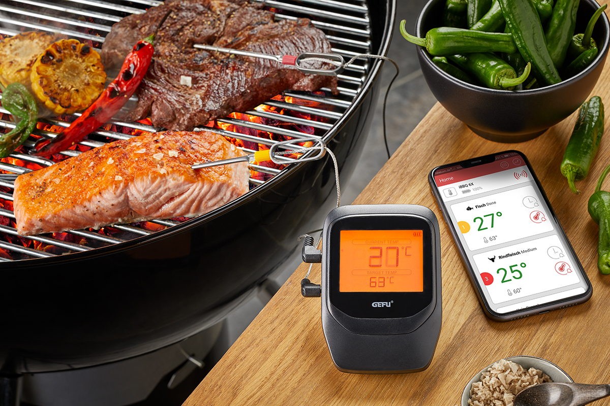 Digitales Fleischthermometer misst die Temperatur von Fleisch auf einem Grill, verbunden mit einer Smartphone-App und umgeben von Paprika.