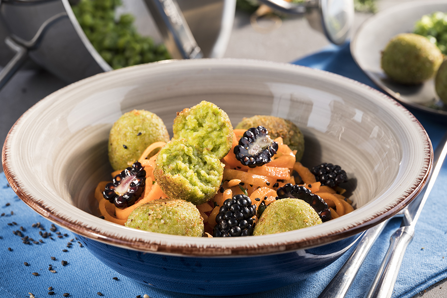 Couscous-Erbsen-Bällchen auf Karotten-Brombeersalat Eine Schale mit grünen Gemüsebällchen, Brombeeren und Möhrennudeln, dekorativ angerichtet auf einem Tisch mit blauer Tischdecke