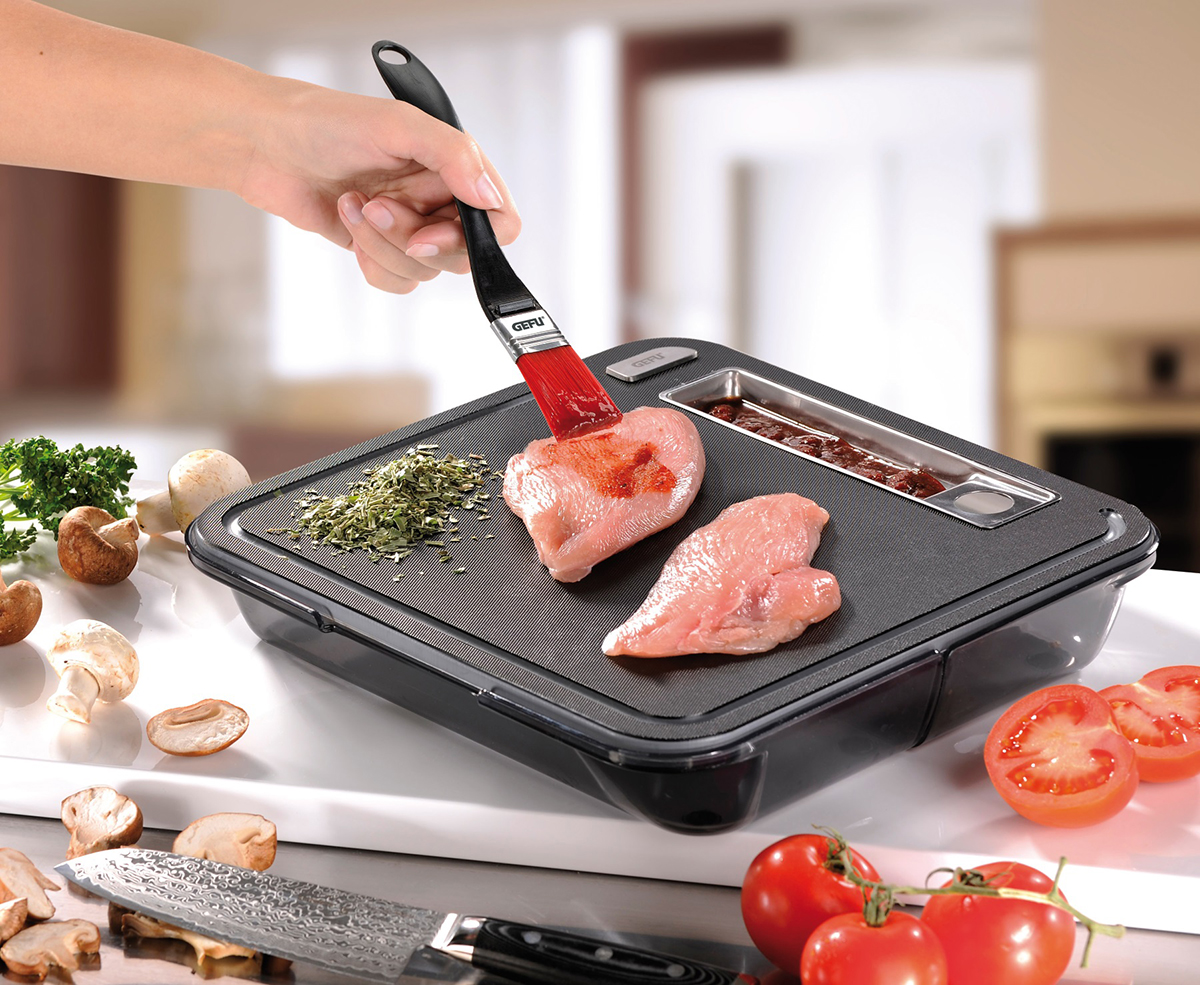 Praktische Küchenarbeit: Würzen von Hähnchenbrust auf Schneidebrett Eine Hand würzt Hähnchenbrust mit einem Pinsel auf einem modernen Schneidebrett mit Kräutern, Gemüse und einem Messer im Hintergrund.