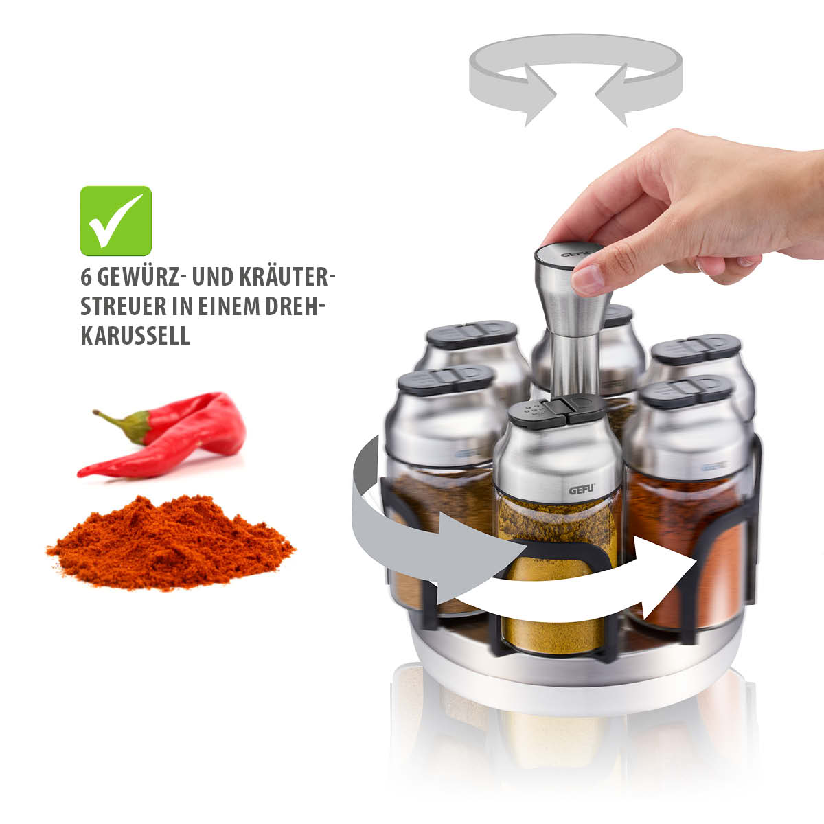 Hand betätigt ein drehbares Gewürzkarussell mit sechs Edelstahlbehältern für Kräuter und Gewürze. Im Hintergrund ist rotes Paprikapulver und eine rote Chili zu sehen.