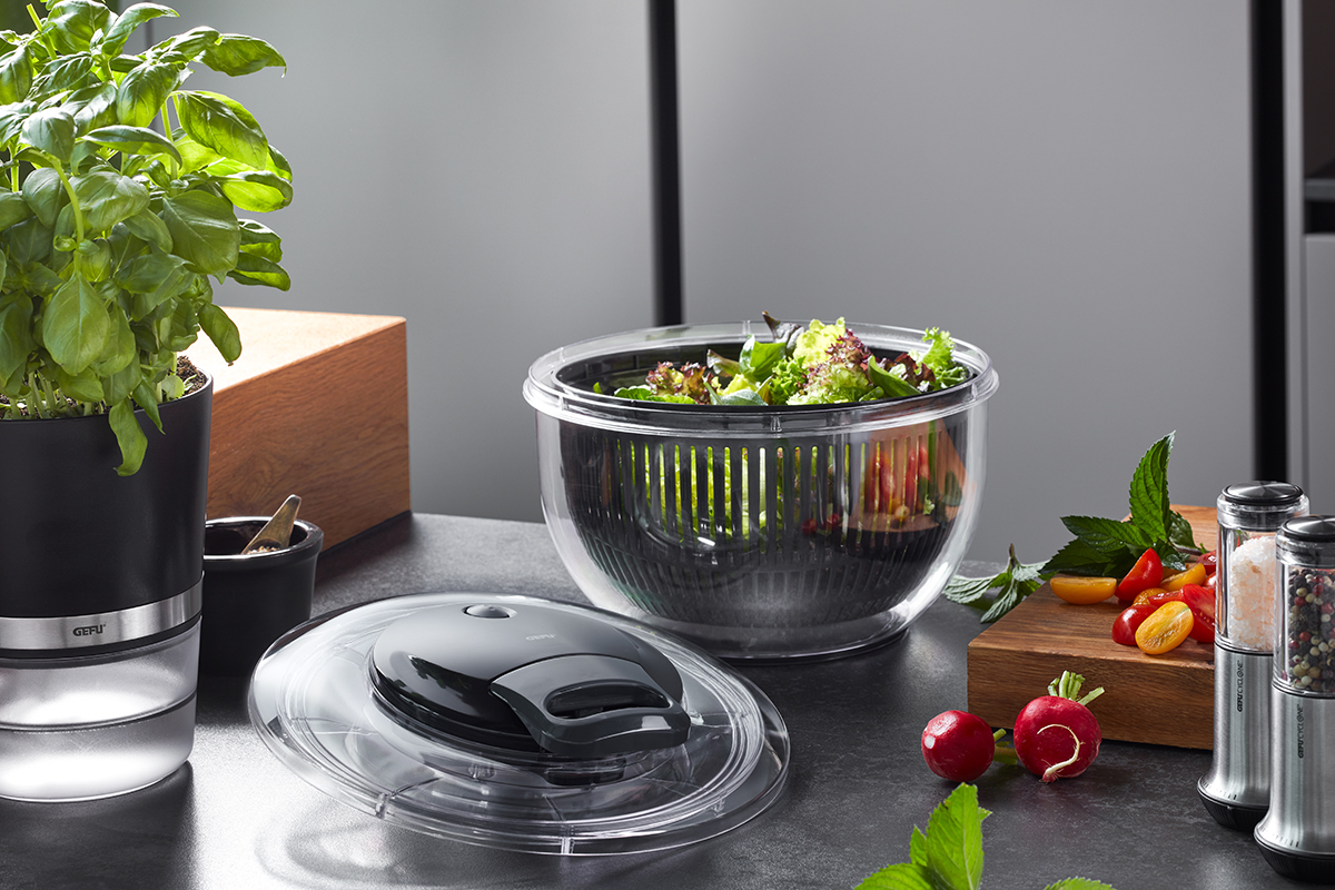 Moderne Küchenszene mit Salatschleuder und frischen Kräutern Küchenarbeitsfläche mit Basilikumpflanze, Salatschleuder voller frischem Salat, Schneidebrett mit frischem Gemüse, Pfeffer- und Salzmühle