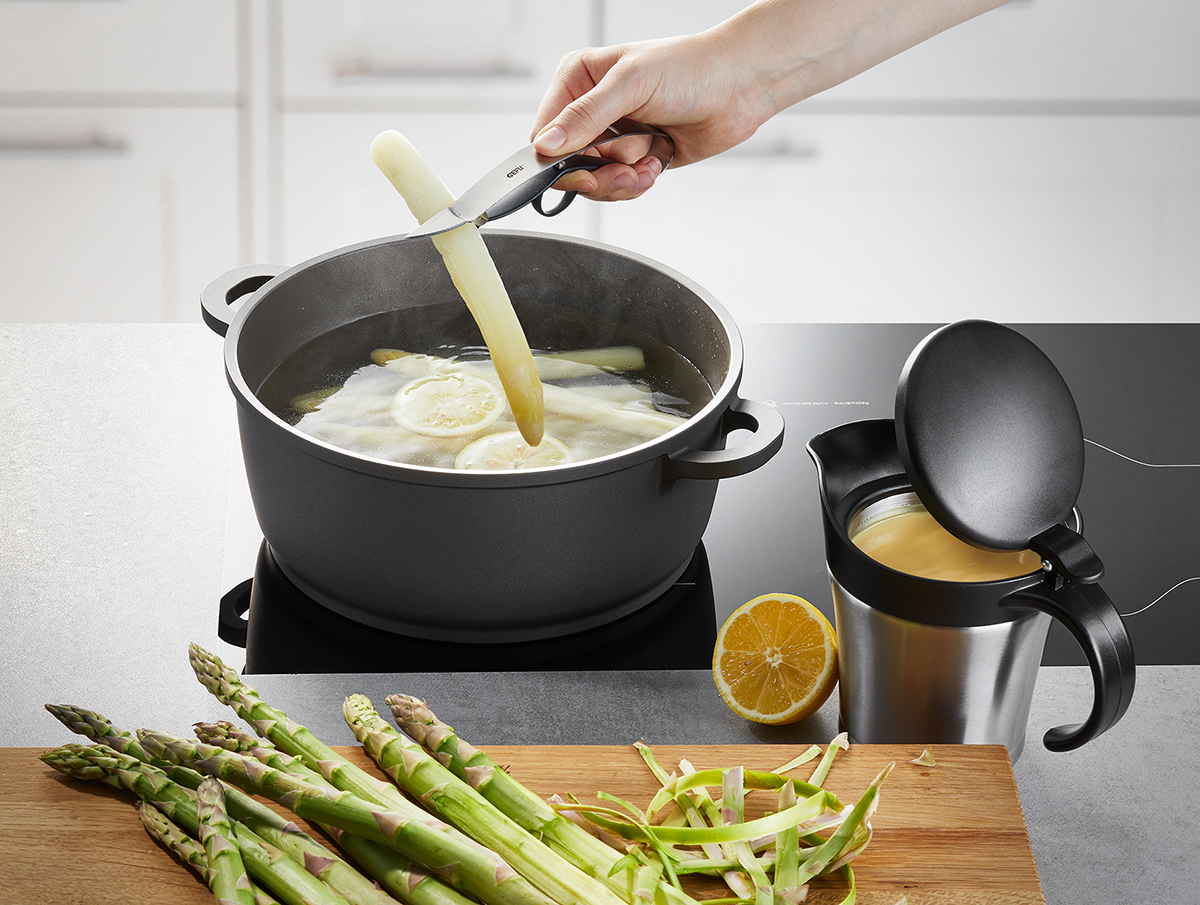 Eine Hand hält Spargel über einem Topf mit kochendem Wasser, neben einer halben Zitrone und einem Behälter mit Sauce. Frischer grüner Spargel liegt auf einem Schneidebrett.