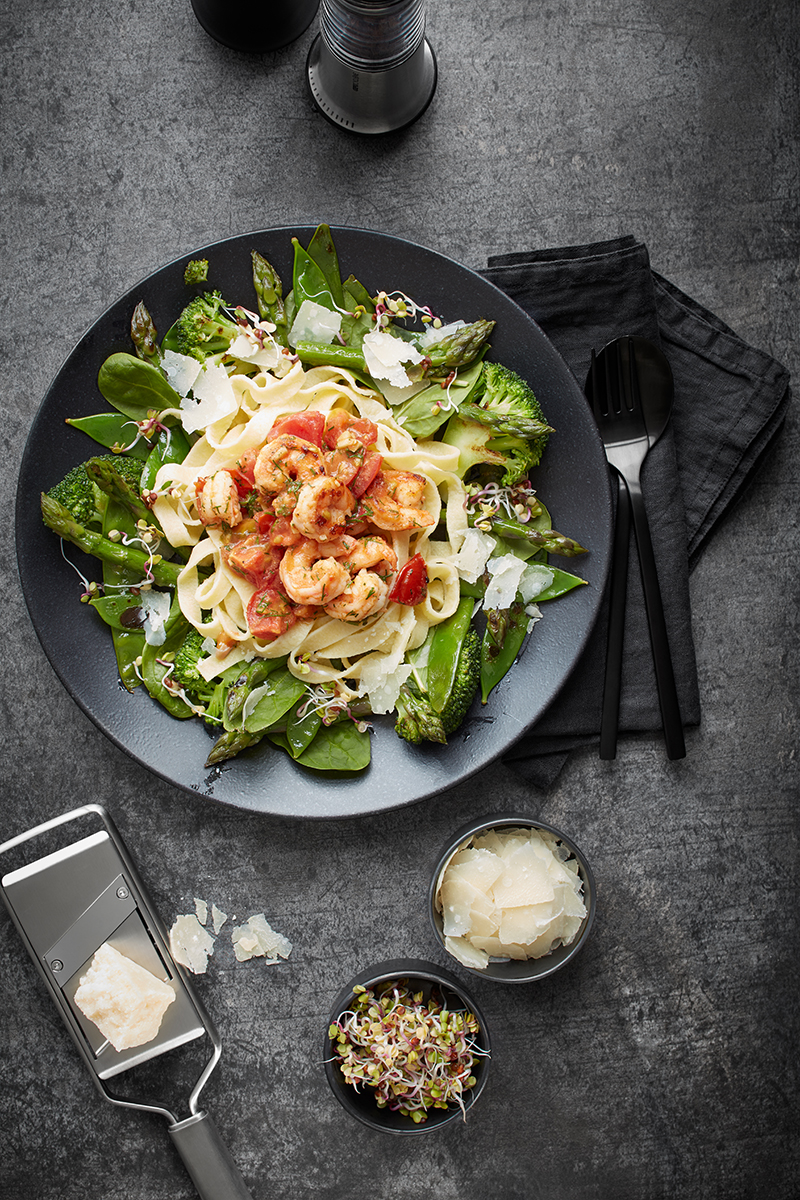 Tagliatelle mit gebratenen Garnelen und Gemüse auf dunklem Teller Ein Teller mit Tagliatelle, frischem Gemüse, gebratenen Garnelen und Parmesan, daneben Pfeffermühle, Besteck und Schalen mit Käse und Sprossen auf grauem Hintergrund