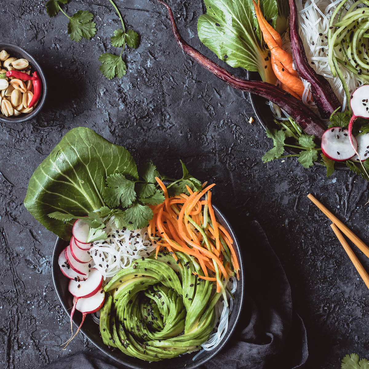 Frischer Salat mit Avocado, Karotten und Radieschen Ein stilvolles Arrangement von frischem Salat mit Avocado, Karotten, Radieschen und Reisnudeln auf einem dunklen Hintergrund, dekoriert mit Koriander und Sesam.