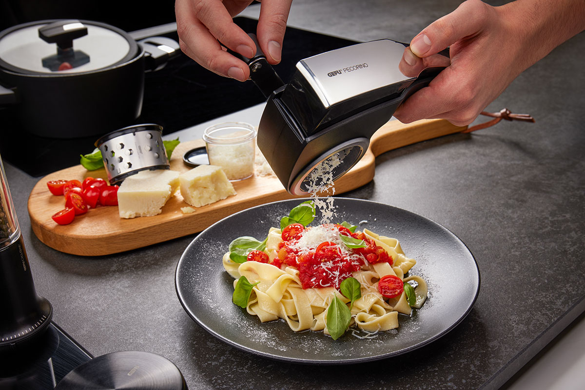 Eine Person reibt frischen Pecorino-Käse auf ein Teller Pasta, garniert mit Tomaten und Basilikumblättern. Im Hintergrund liegt eine Schneidebrett mit Käse, Tomaten und einer Käsereibe.