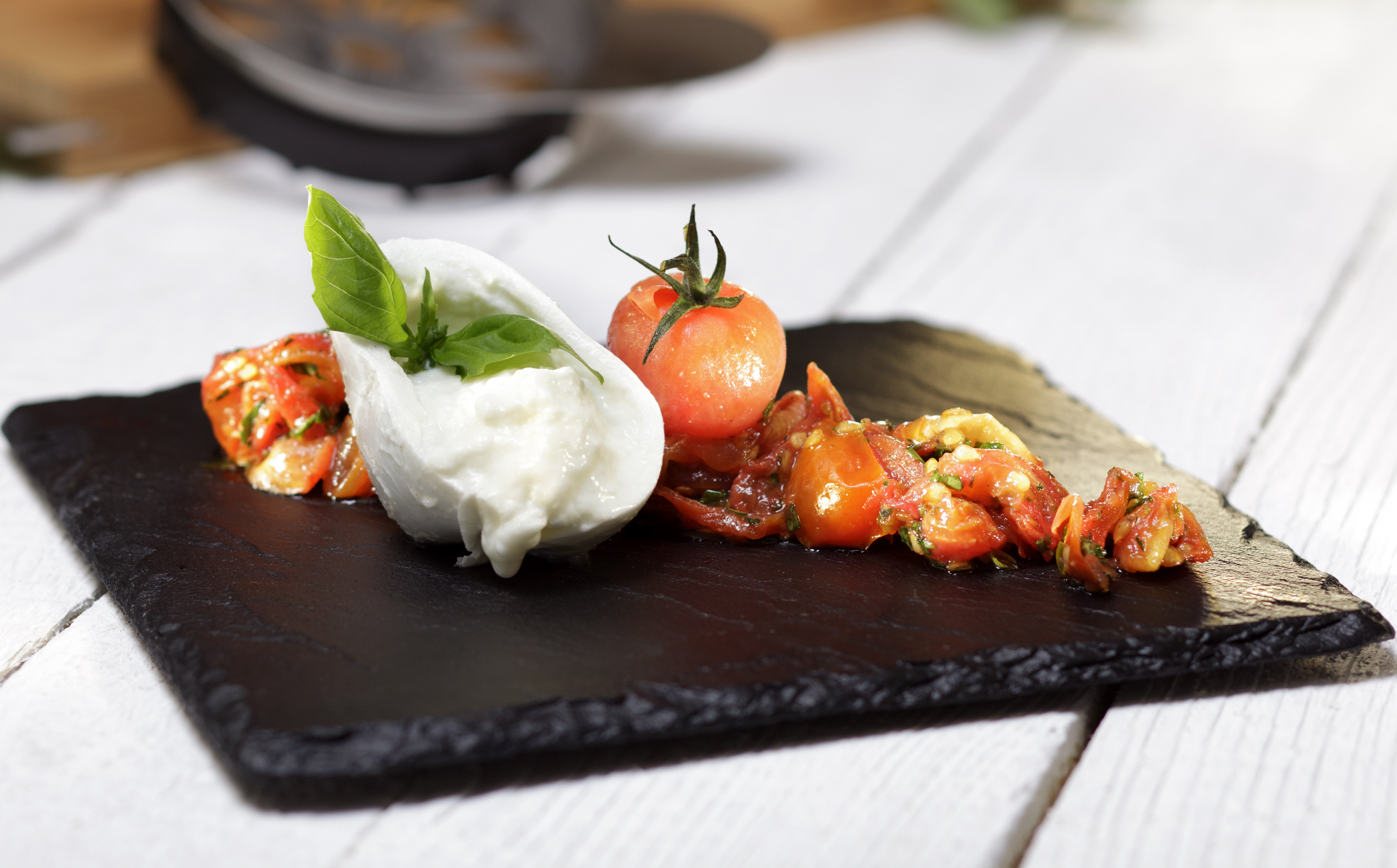 Italienischer Burrata-Käse mit gesüßten Tomaten  Frische Burrata mit Basilikum und Tomaten auf einer eleganten Schieferplatte, appetitlich angerichtet für ein Gourmet-Erlebnis