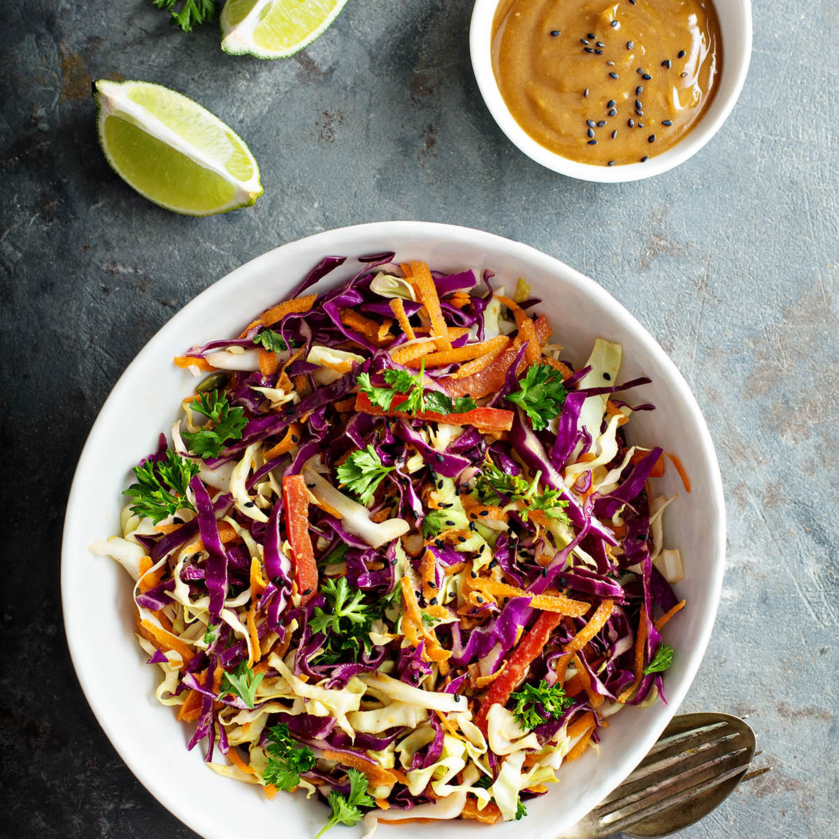 Frischer Krautsalat mit Karotten und Petersilie, serviert mit einer Erdnusssauce Bunte Salatschüssel mit Weiß- und Rotkohl, Karotten und Kräutern, neben Limetten und einer Schüssel Erdnusssauce