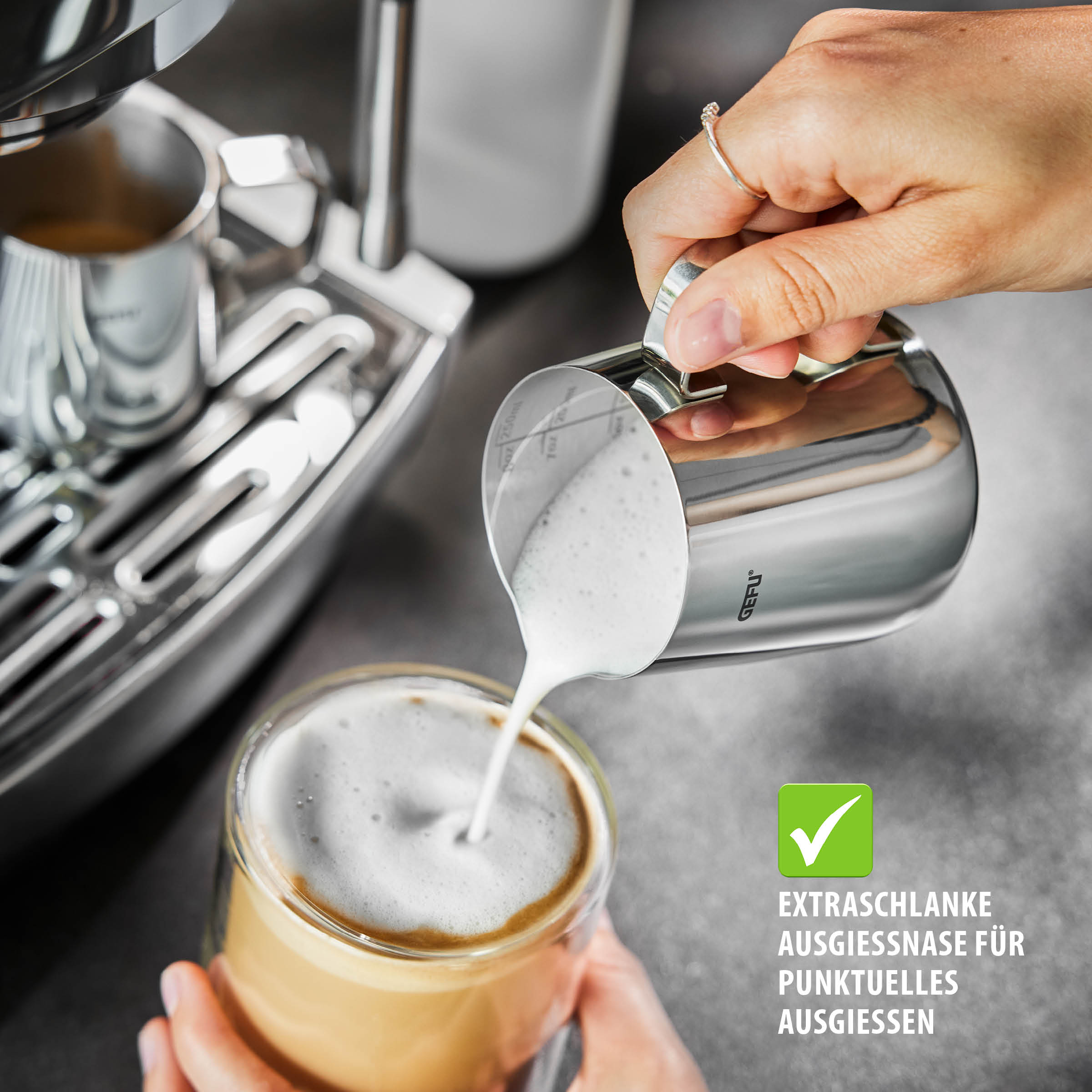 Milchkännchen BARISTA 250 ml von GEFU aus Edelstahl mit Skalierung und extraschlanker Ausgießnase, beim punktgenauen Eingießen von Milchschaum in einen Kaffee neben einer Espressomaschine.