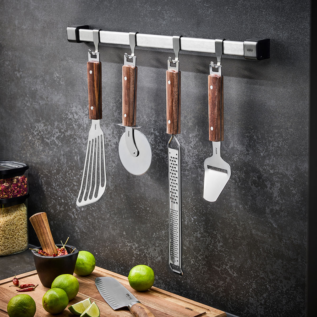 Moderne Küchenutensilien mit Holzgriff an Hakenleiste Küchenutensilien wie Pfannenwender und Pizzaschneider mit Holzgriff an Wandhaken, daneben Limonen auf Schneidebrett