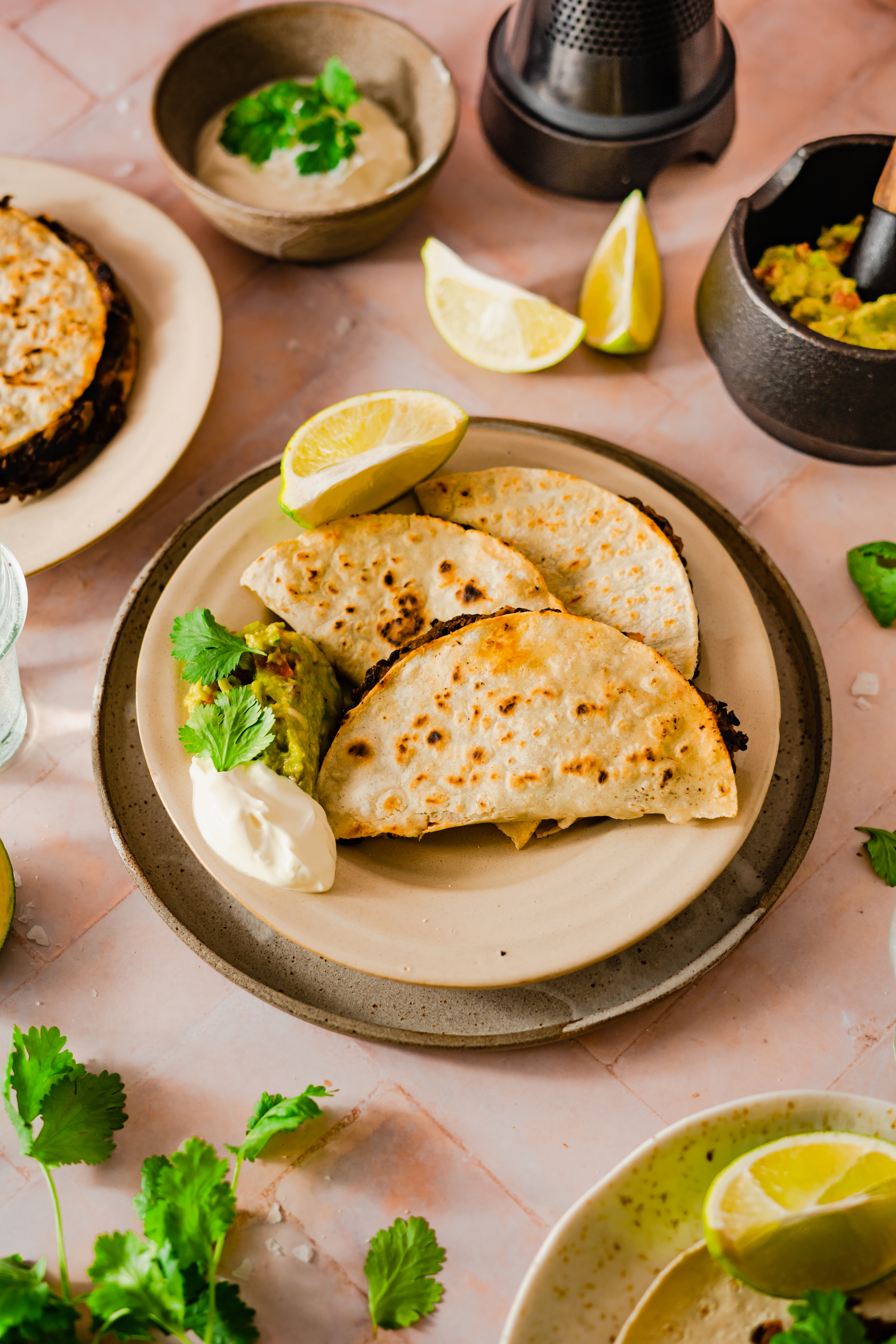 Schwarze Bohnen Tacos mit Guacamole Ein Teller mit Quesadillas, serviert mit Guacamole, Sauerrahm und Limettenscheiben, umgeben von frischen Kräutern.