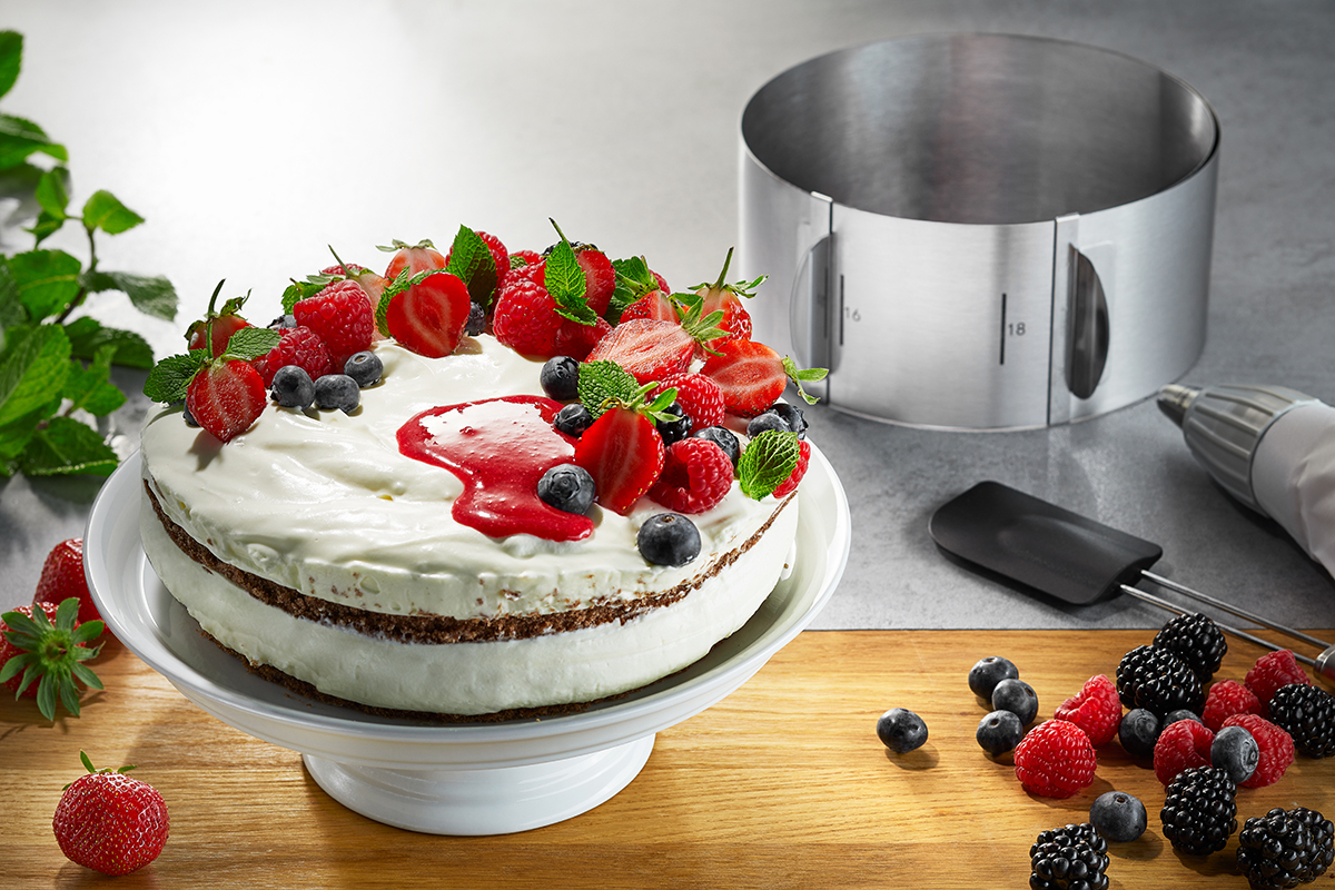 Kuchen mit Beeren und Sahne auf Tortenplatte Ein dekorierter Kuchen mit verschiedenen Beeren und Sahne auf einer weißen Tortenplatte, daneben ein Tortenring und Backutensilien.