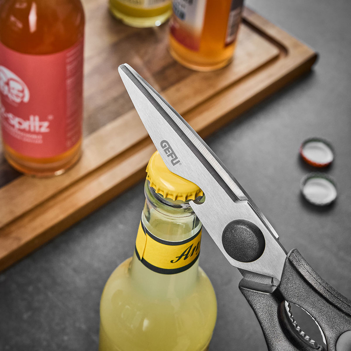 Eine ine Schere öffnet den Kronkorken einer Flasche, im Hintergrund weitere Getränke.