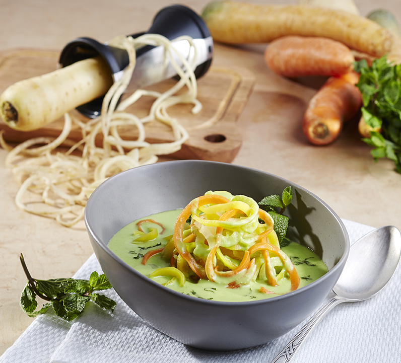 Frühlingseintopf mit Kokosmilch und Minze Ein Teller mit Gemüsenudeln in grüner Kräutersauce, daneben ein Spiralschneider und frische Zutaten auf einem Tisch