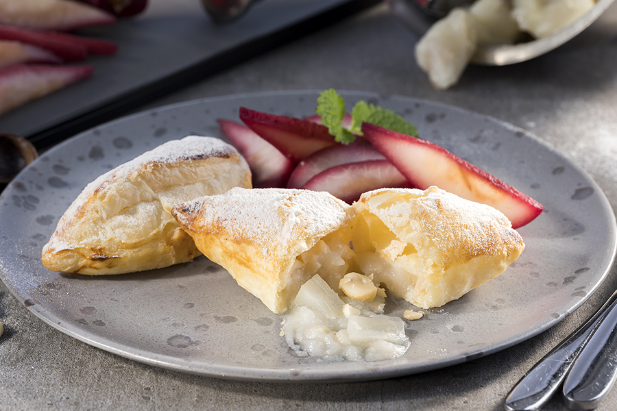 Puff pastry pockets with pear-macadamia filling and pickled red wine pears Ein Dessert aus Blätterteig, gefüllt mit Creme, serviert mit frischen Birnenscheiben auf einem grauen Teller