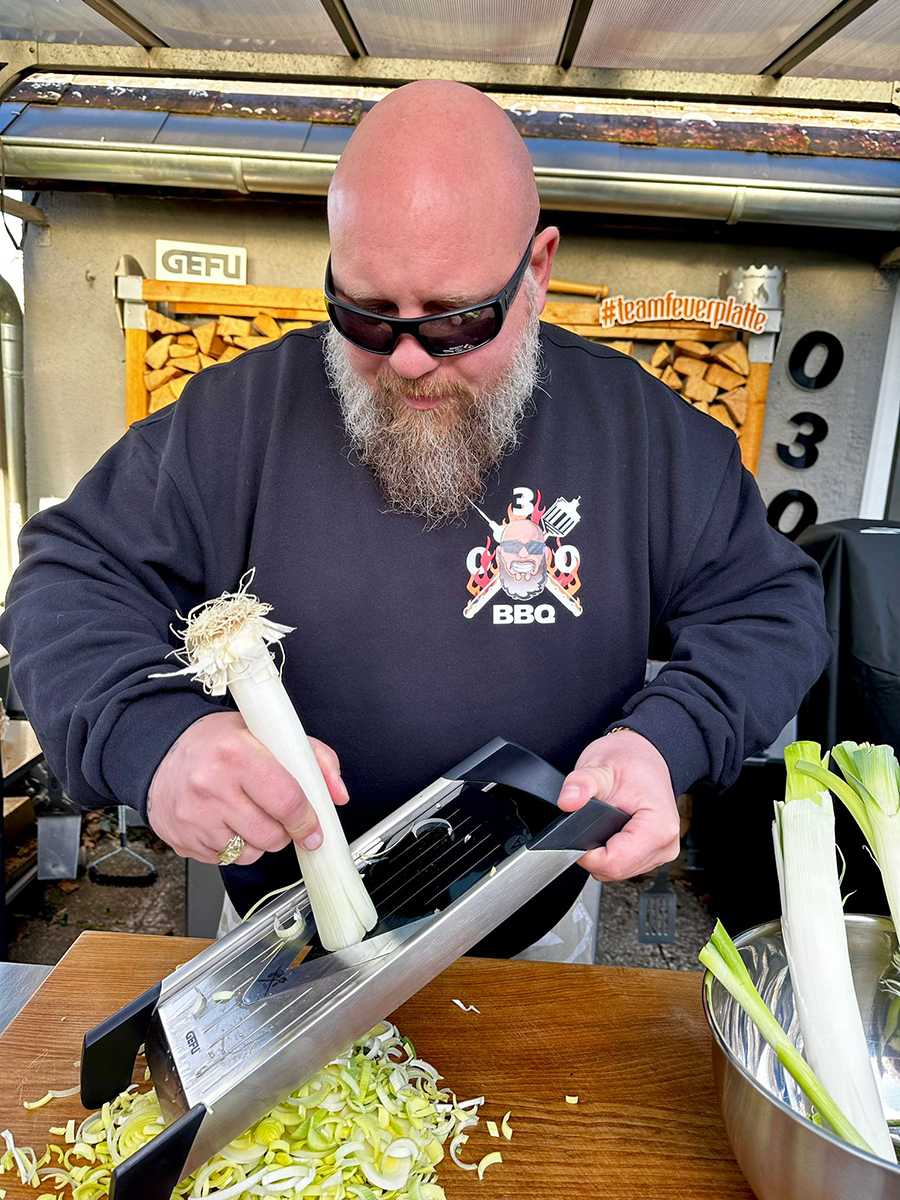 Person schneidet Lauch mit Hobel in der Küche Eine Person mit Sonnenbrille schneidet frischen Lauch auf einem Holzbrett mit einem Gemüsehobel.