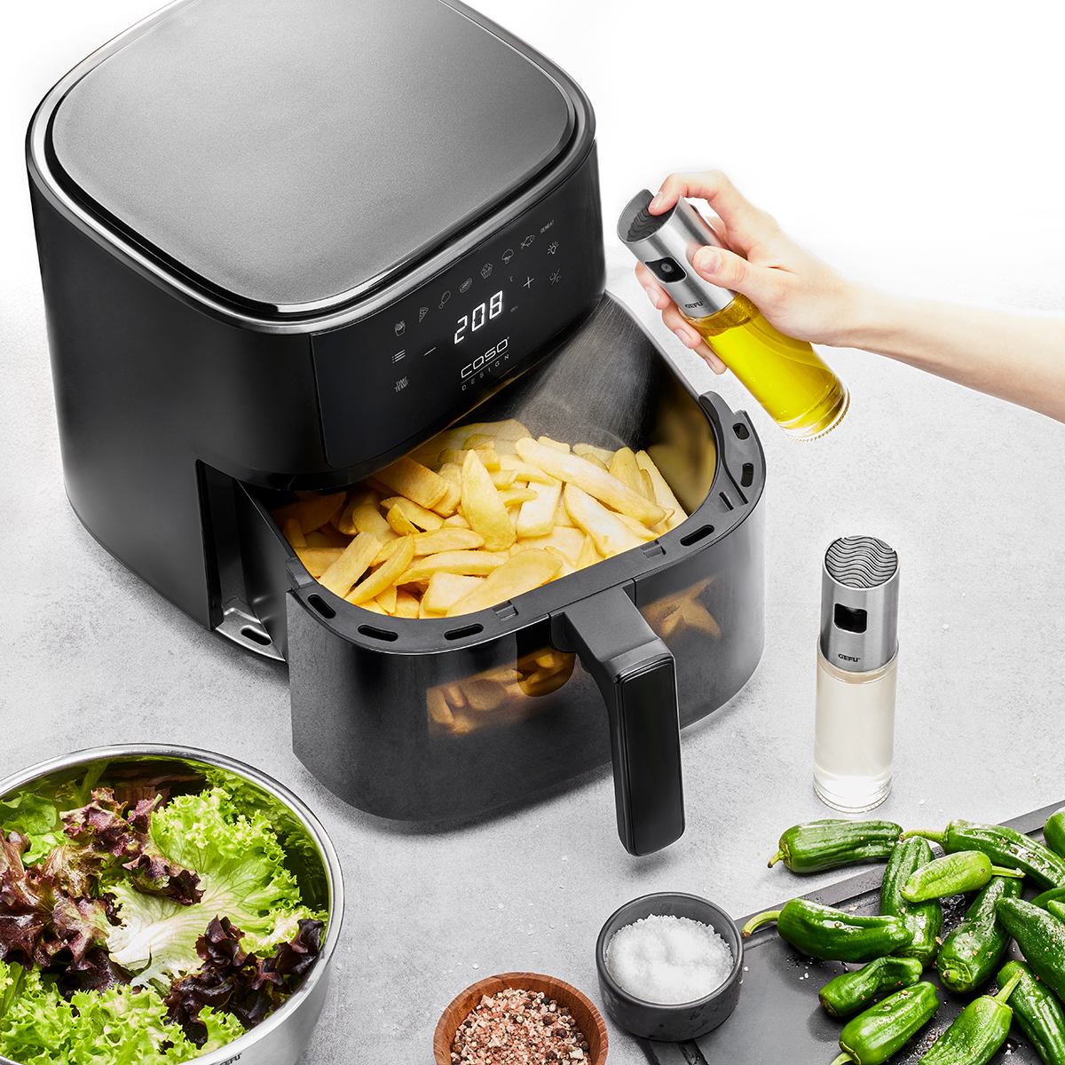 Eine Heißluftfritteuse mit einer geöffneten Schublade voller Pommes frites, ein Ölsprüher wird darüber verwendet. Daneben liegen Salat, Gewürze und grüne Paprikaschoten auf einem grauen Küchentisch.
