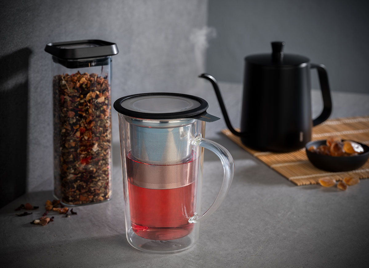 Doppelwandiger Teebecher mit Teesieb und Teekanne im Hintergrund Ein doppelwandiger Glas-Teebecher mit darin befindlichem Teesieb, gefüllt mit rotem Tee, steht neben einem Glasbehälter mit losen Teeblättern und einer schwarzen Teekanne.