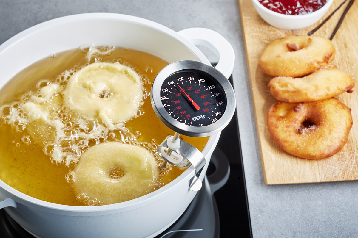 Frittieren von Apfelringen in heißem Öl mit Küchenthermometer Apfelringe werden in heißem Öl in einem weißen Topf frittiert, überwacht von einem Küchenthermometer, daneben fertige Apfelringe auf einem Holzbrett mit Marmelade.
