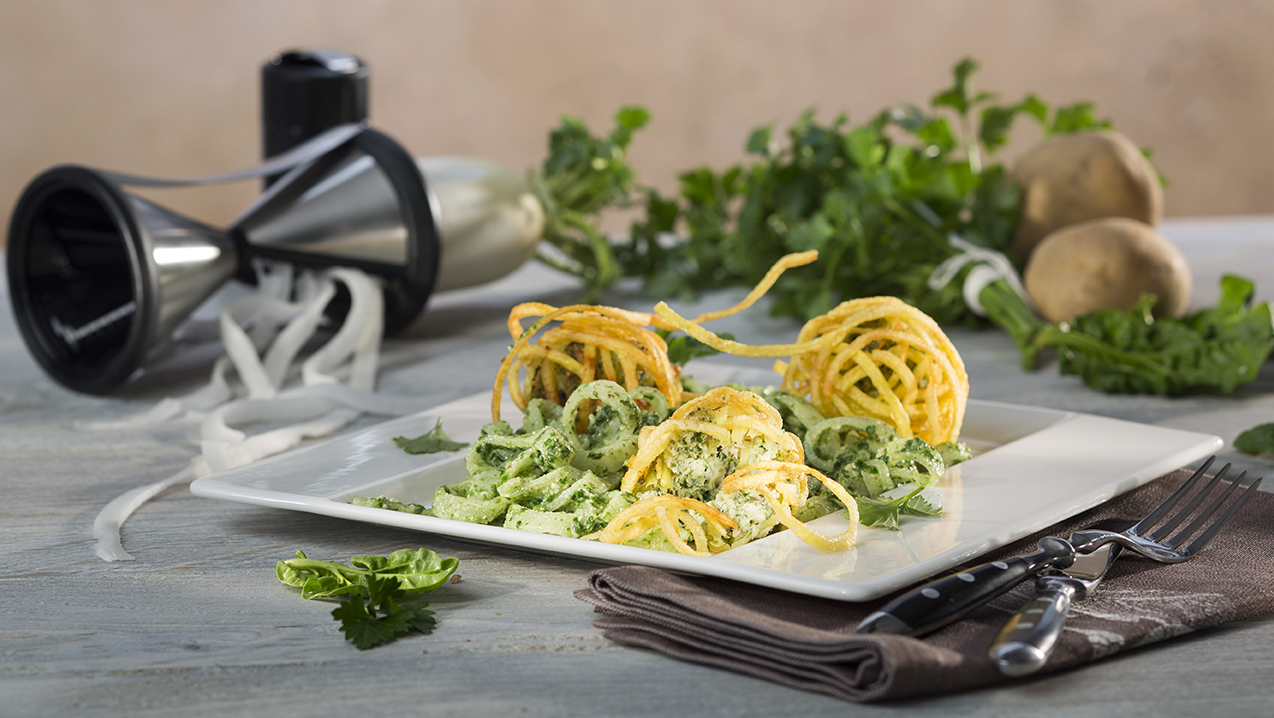 Trout croustillons on radish salad with parsley pesto Ein Teller mit Zucchininudeln, garniert mit Kartoffelspiralen und frischen Kräutern. Im Hintergrund ein Gemüseschneider und frische Zutaten.