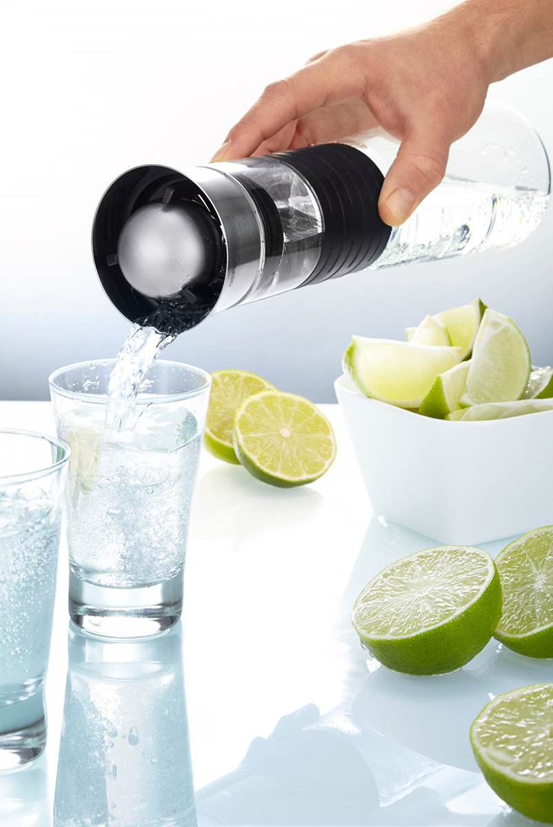 Elegante Wasserkaraffe mit Eisfilter beim Eingießen von erfrischendem Wasser Hand gießt Wasser aus einer Karaffe mit Eisfilter in ein Glas, umgeben von frischen Limettenscheiben und Schale