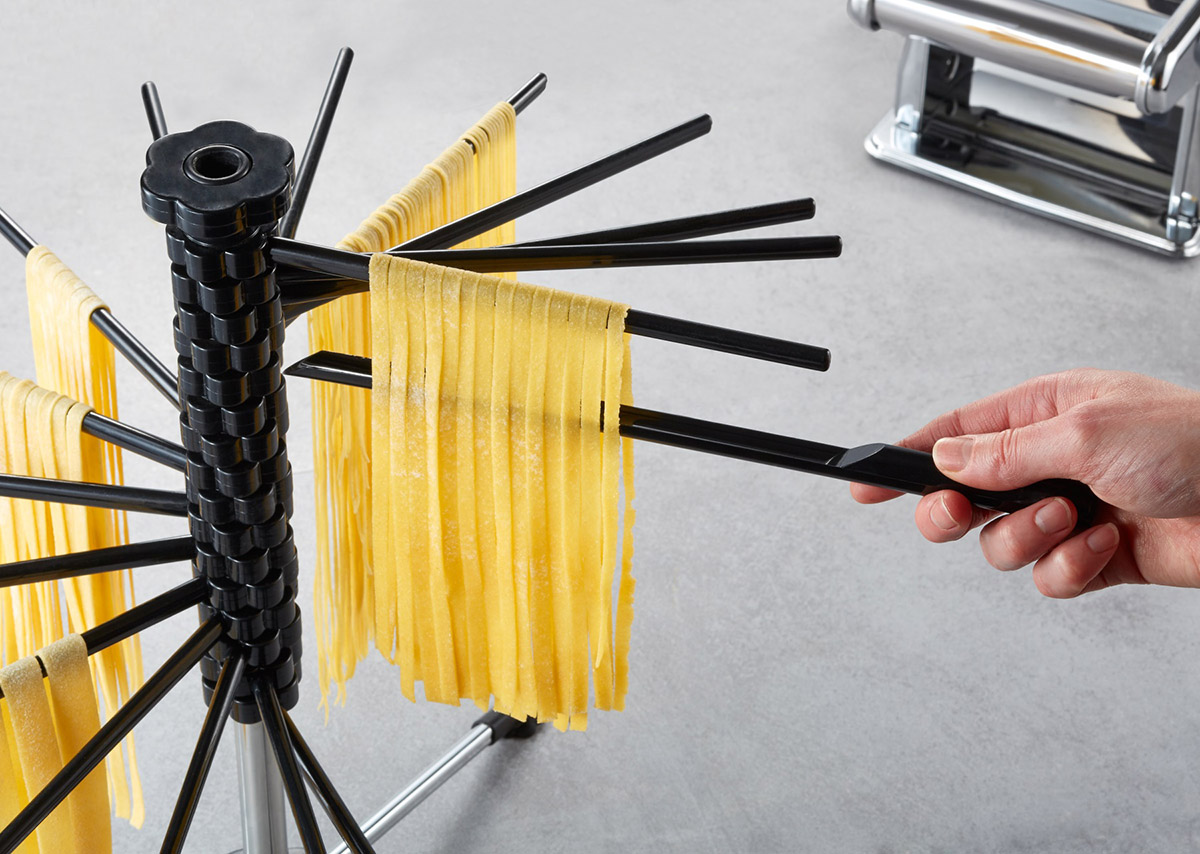 Nudeltrockner mit frischer Pasta Ein schwarzer Nudeltrockner mit mehreren Armen, auf denen frische Nudeln zum Trocknen ausgelegt sind. Im Hintergrund ist eine Nudelmaschine sichtbar.