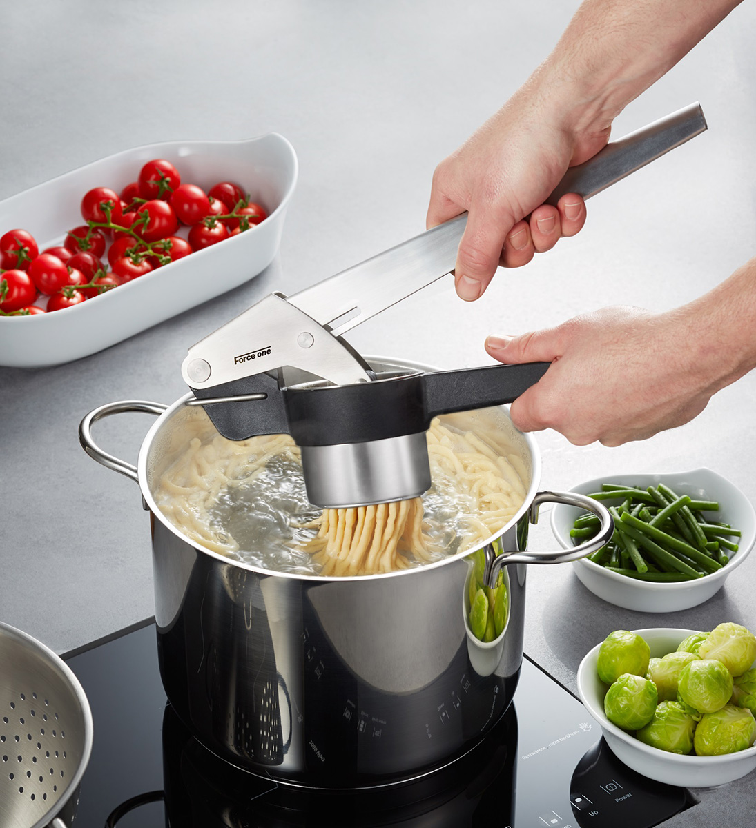 Manuelle Spätzlepresse über Kochtopf mit frischen Zutaten Eine Hand hält eine Spätzlepresse über einem Kochtopf auf einem Herd. Im Hintergrund sind Schalen mit Tomaten, Bohnen und Rosenkohl zu sehen.