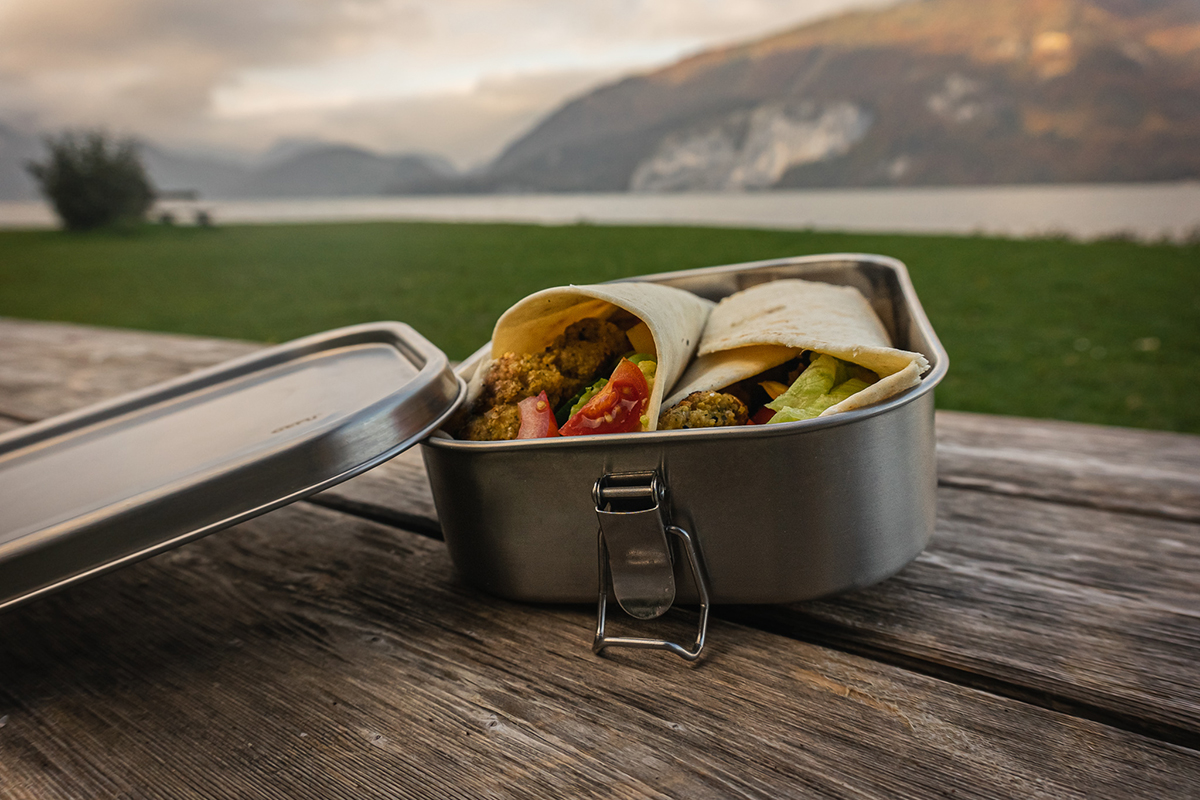 Eine geöffnete Edelstahl-Brotbox mit zwei frischen Wraps, gefüllt mit Salat und Tomaten, auf einem Holztisch vor einer malerischen Seelandschaft.