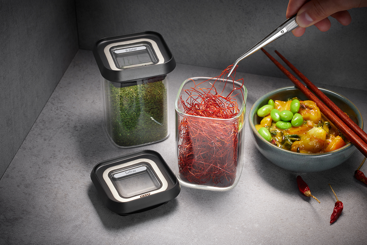 Gewürzgläser mit Kräutern und Safran neben einer Schale asiatischer Speisen Zwei Vorratsgläser mit Kräutern und Safran, eine Schale mit asiatischem Gericht und Essstäbchen, auf einer grauen Arbeitsfläche