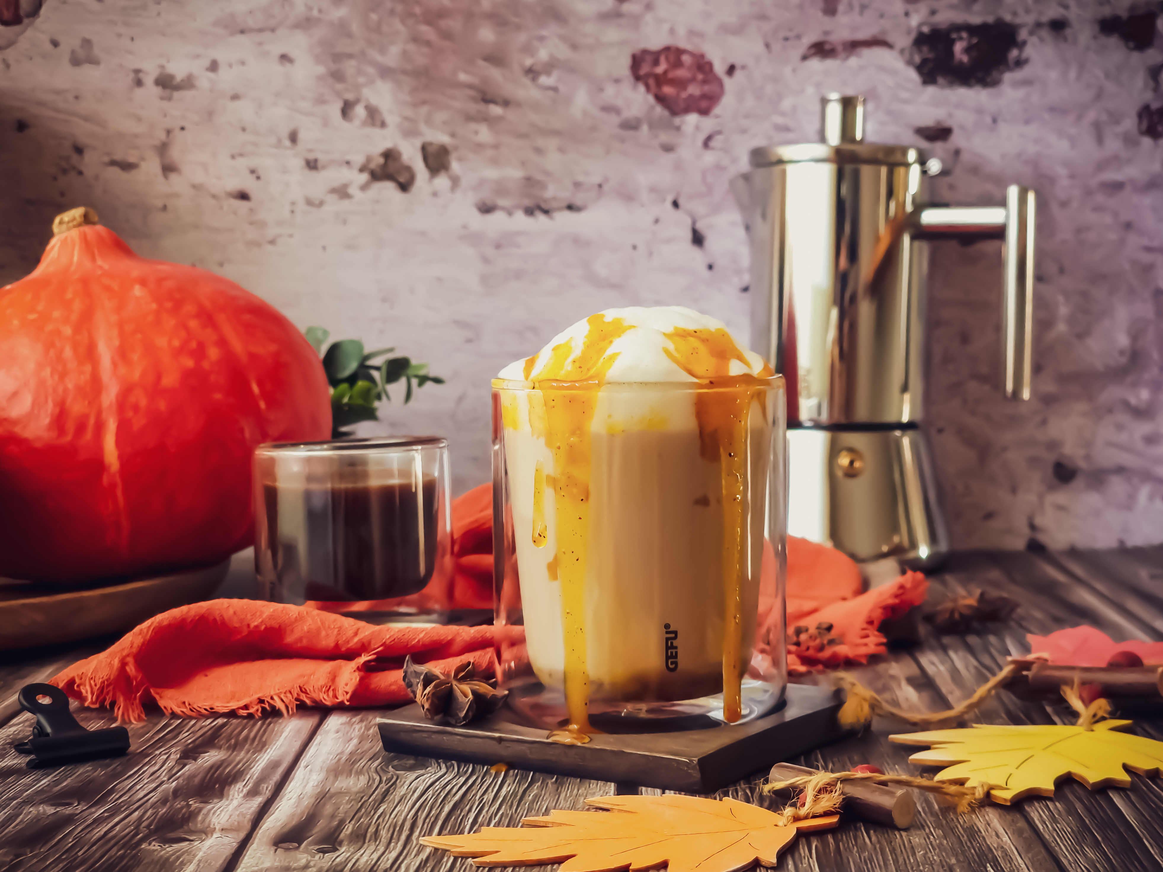 Pumpkin Latte  Ein Glas Kürbis-Latte mit Sahne und Karamell neben einem Orange Kürbis und einer silbernen Espressokanne auf einem Holztisch mit Herbstdekoration.