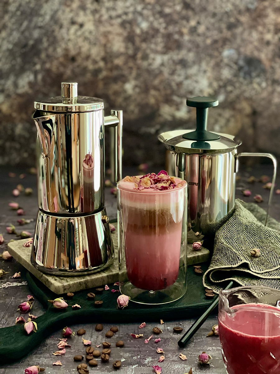 Elegante Espressokanne und Glas mit rosa Kaffee Eine glänzende Espressokanne neben einem Glas mit rosa Kaffee und Rosenblüten, umgeben von Kaffeebohnen auf einem rustikalen Tisch