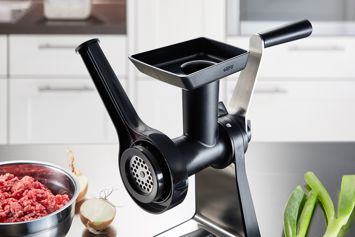 Hochwertiger Fleischwolf in moderner Küche Ein schwarzer Fleischwolf auf einer Küchenarbeitsplatte mit Schüssel Hackfleisch, Zwiebeln und Frühlingszwiebeln im Hintergrund.