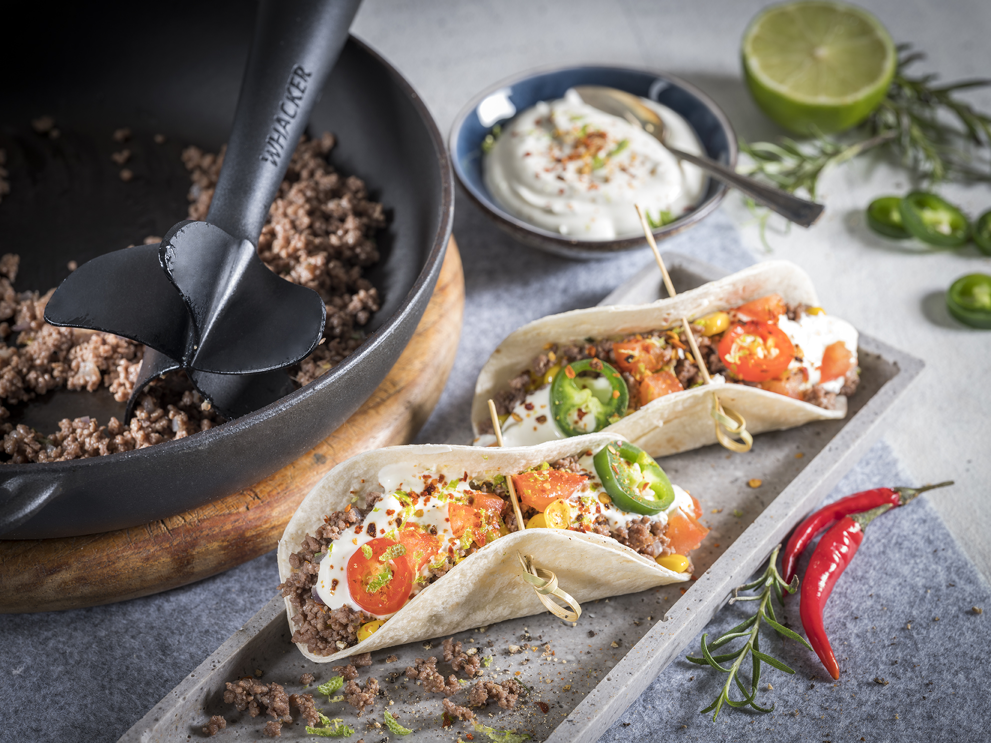 Mexikanische Hackfleisch-Chili-Tacos  Zwei Tacos gefüllt mit Hackfleisch, Tomaten, Jalapeños, Joghurtsauce und Gewürzen auf einem Tablett neben einer Pfanne mit Hackfleisch und einer Limette
