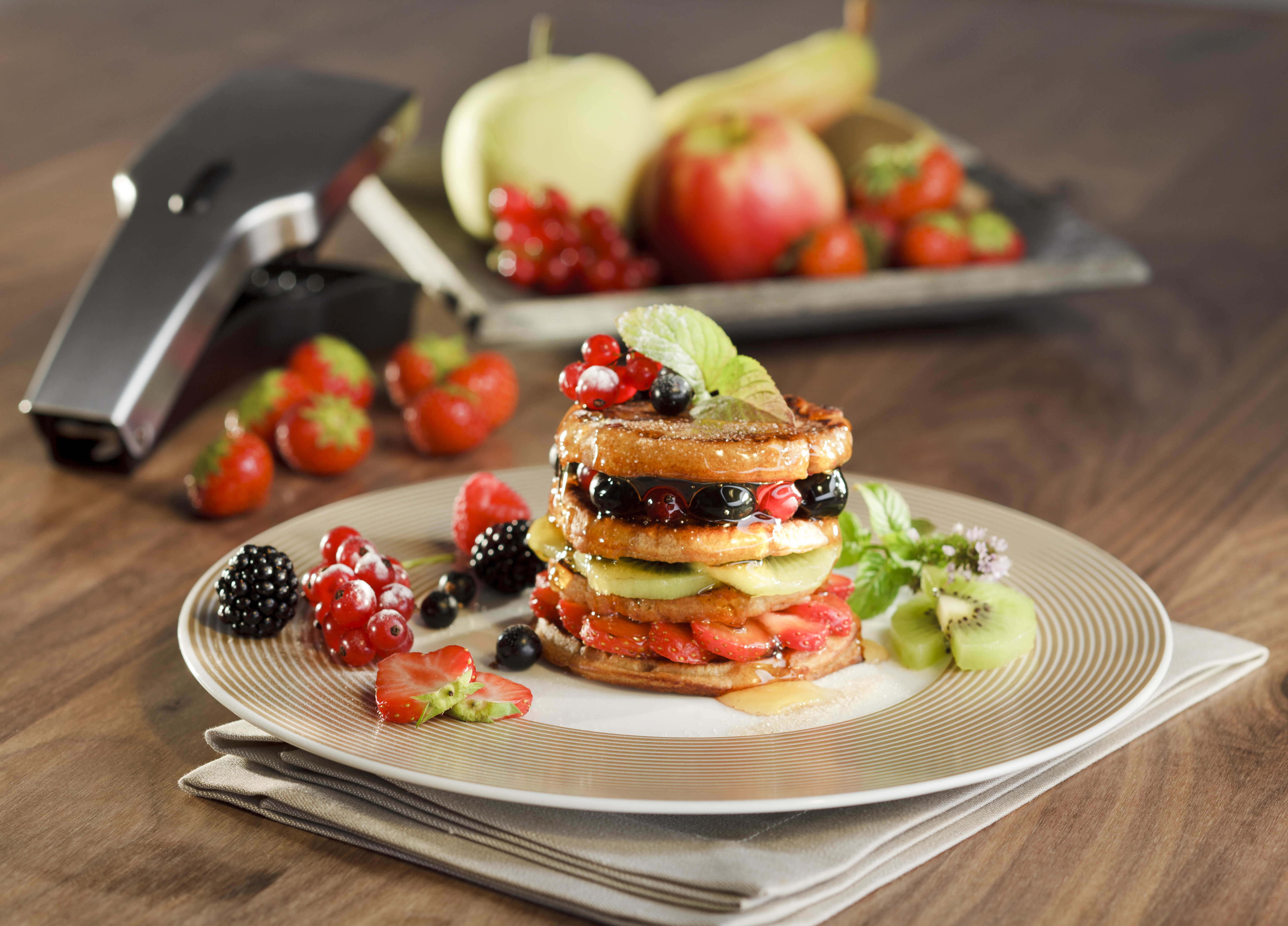 Sweet pancake fruit tower Ein Pfannkuchenturm garniert mit frischen Beeren, Fruchtscheiben und Minzblättern auf einem Teller, umgeben von Erdbeeren und Äpfeln im Hintergrund.