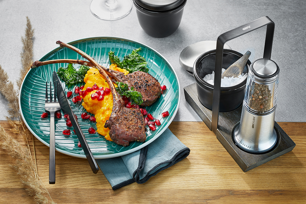 Gegrillte Lammkoteletts mit Kürbispüree und Granatapfelkernen auf einem grünen Teller, serviert mit Salz und Pfeffermühle Ein elegantes Gericht aus Lammkoteletts, Kürbispüree und Granatapfel, begleitet von einer Salz- und Pfeffermühle auf einem Holzbrett