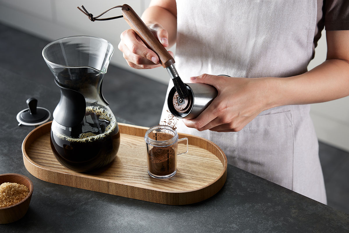 Handkaffeemühle mit frisch gebrühtem Kaffee auf Holztablett Eine Person mahlt Kaffee mit einer Handkaffeemühle neben einer Karaffe frisch gebrühten Kaffees auf einem Holztablett.