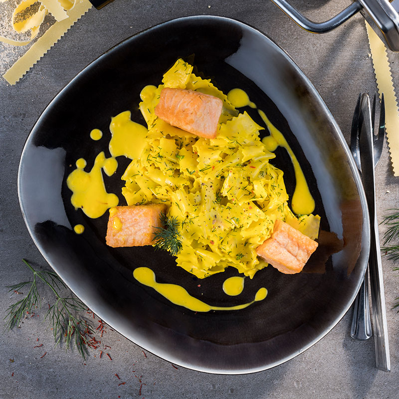Elegantes Pastagericht mit Lachs und gelber Sauce auf schwarzem Teller Nahaufnahme eines modernen Pastagerichts mit Lachs und gelber Sauce, serviert auf einem schwarzen Teller