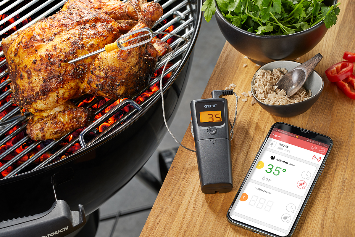 Ein gegrilltes Hühnchen auf einem Grill, überwacht durch ein digitales Thermometer und eine Smartphone-App, um die richtige Temperatur zu gewährleisten.