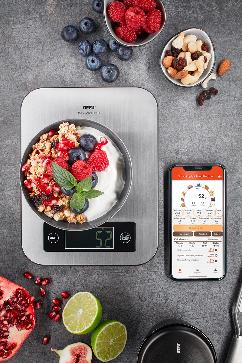 Digitale Küchenwaage mit gesunder Himbeer-Joghurt-Schüssel und Smartphone-App Digitale Küchenwaage zeigt Gewicht einer Schüssel mit Joghurt, Müsli, Beeren und Minzblatt neben einem Smartphone mit Ernährungs-App.