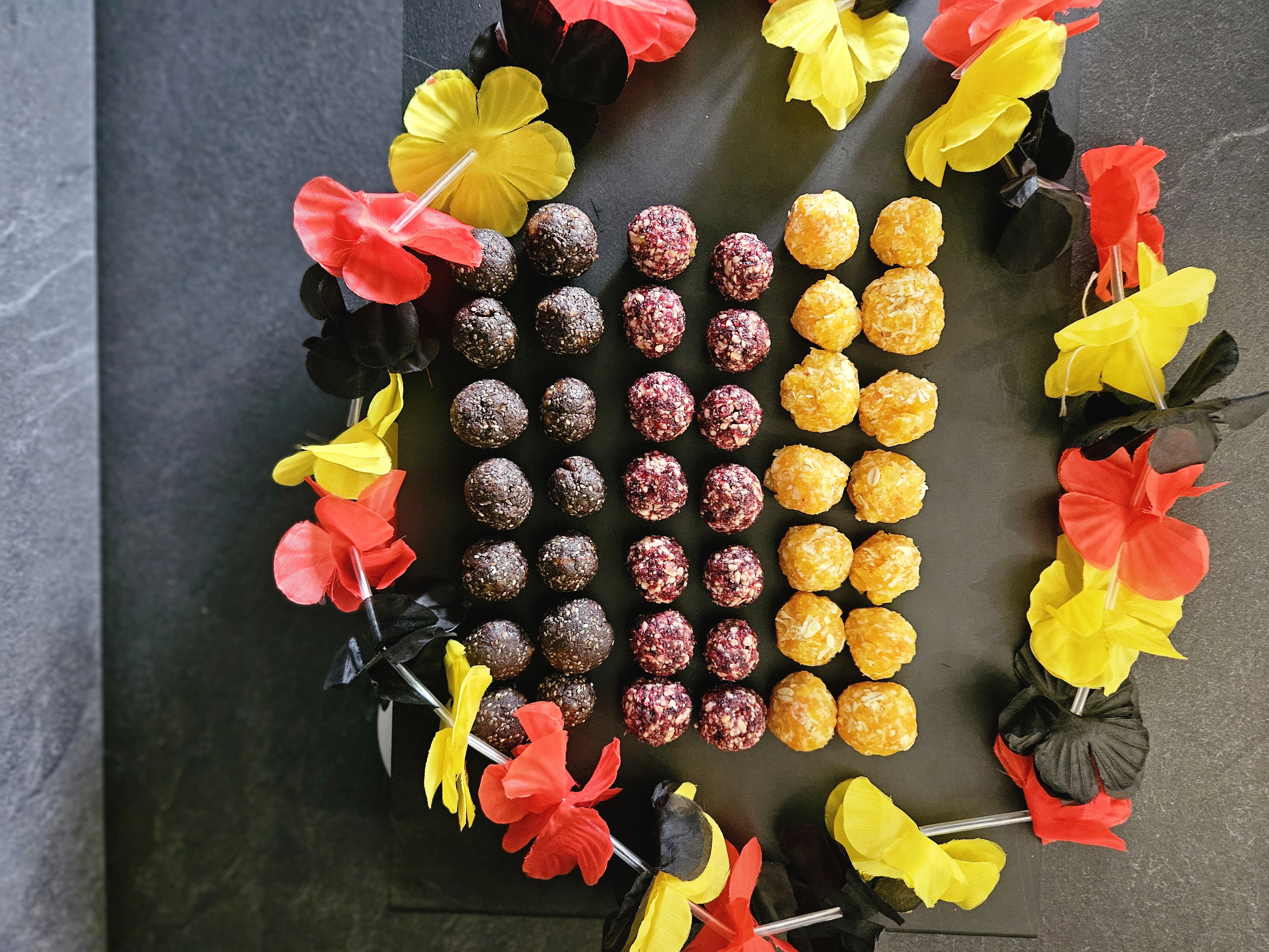 EM Energy Balls Verschiedene Pralinen auf einer schwarzen Platte, umrandet von roten, gelben und schwarzen Blumen