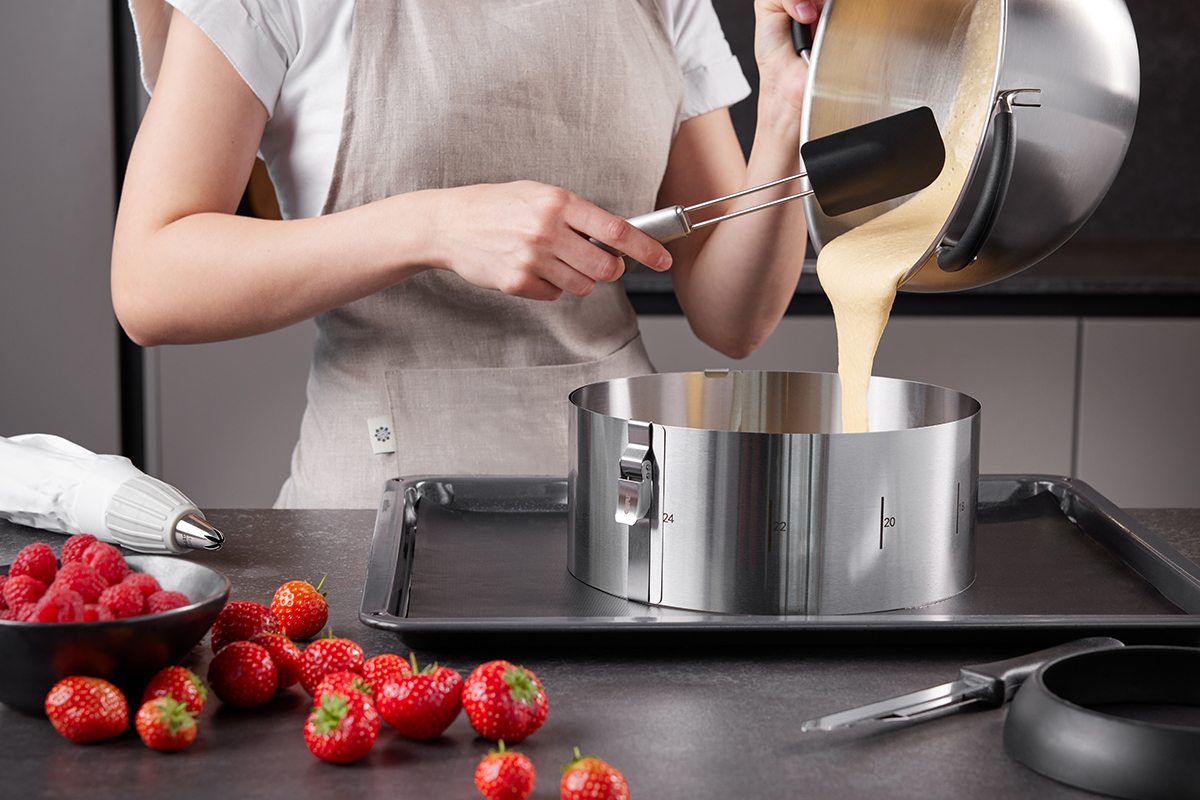 Person füllt Kuchenteig in eine runde Backform Person gießt flüssigen Teig aus einem Topf in eine runde Backform auf einem Backblech, umgeben von Erdbeeren und Himbeeren