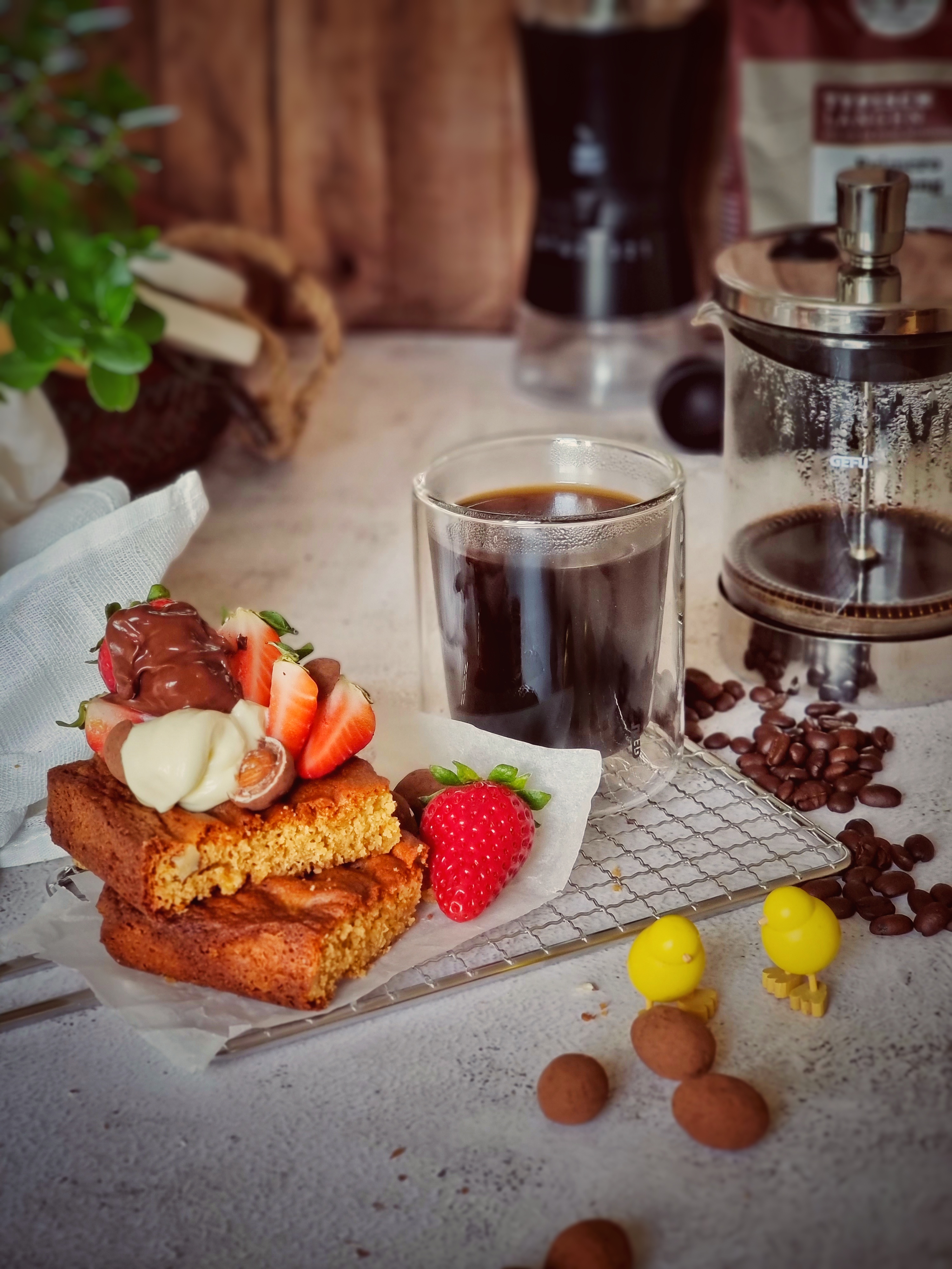 Erdbeerbrownie  Ein Glas schwarzer Kaffee neben einer Französischpresse, umgeben von Kuchen mit Erdbeeren und Sahne sowie Kaffeebohnen auf einem Tisch.