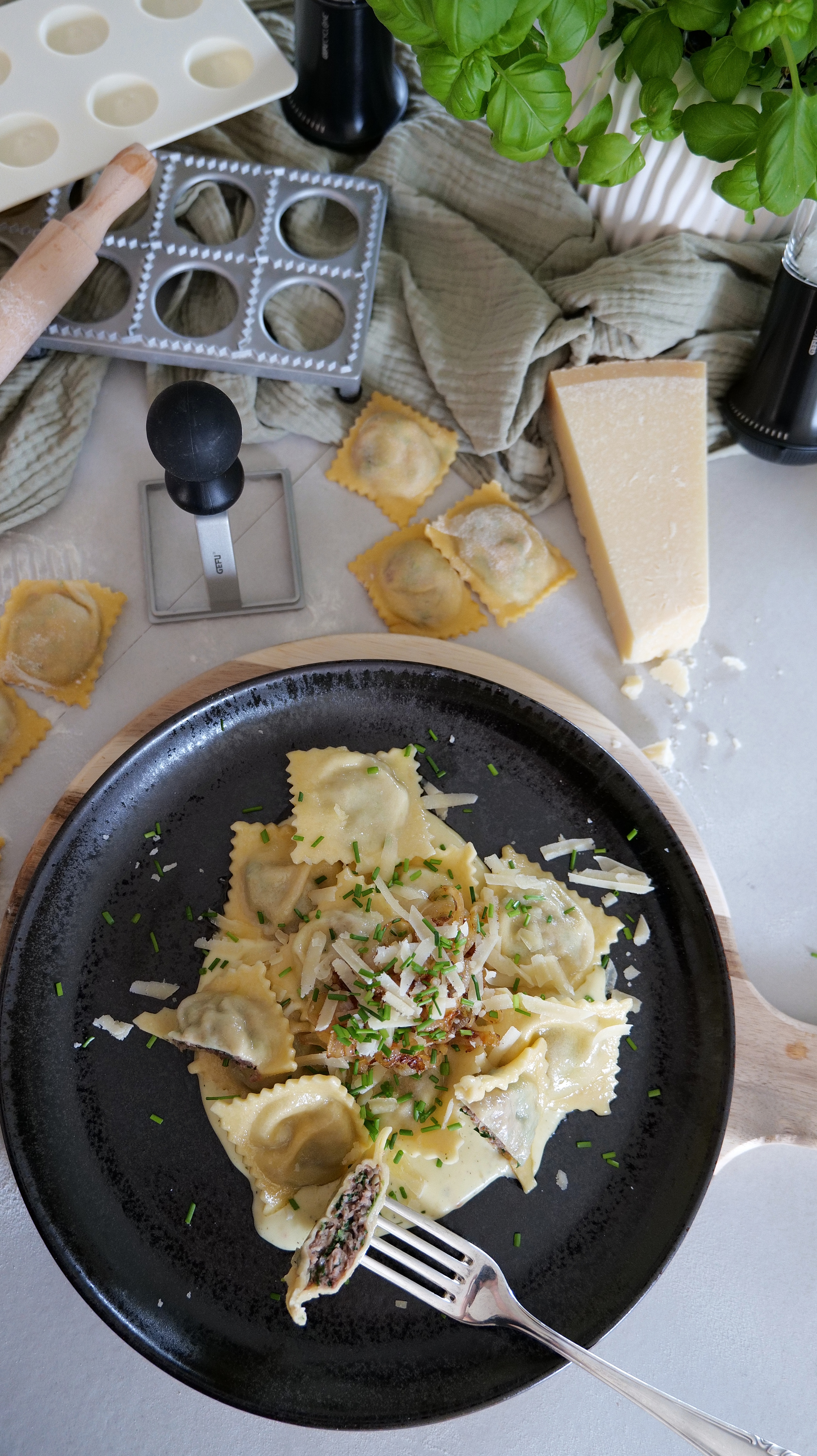 Ravioli im Maultaschenstyle Ein schwarzer Teller mit hausgemachten Ravioli, garniert mit geriebenem Parmesan und frischen Kräutern. Daneben liegen Ravioli-Werkzeuge und ein Stück Parmesankäse auf einem Tisch.