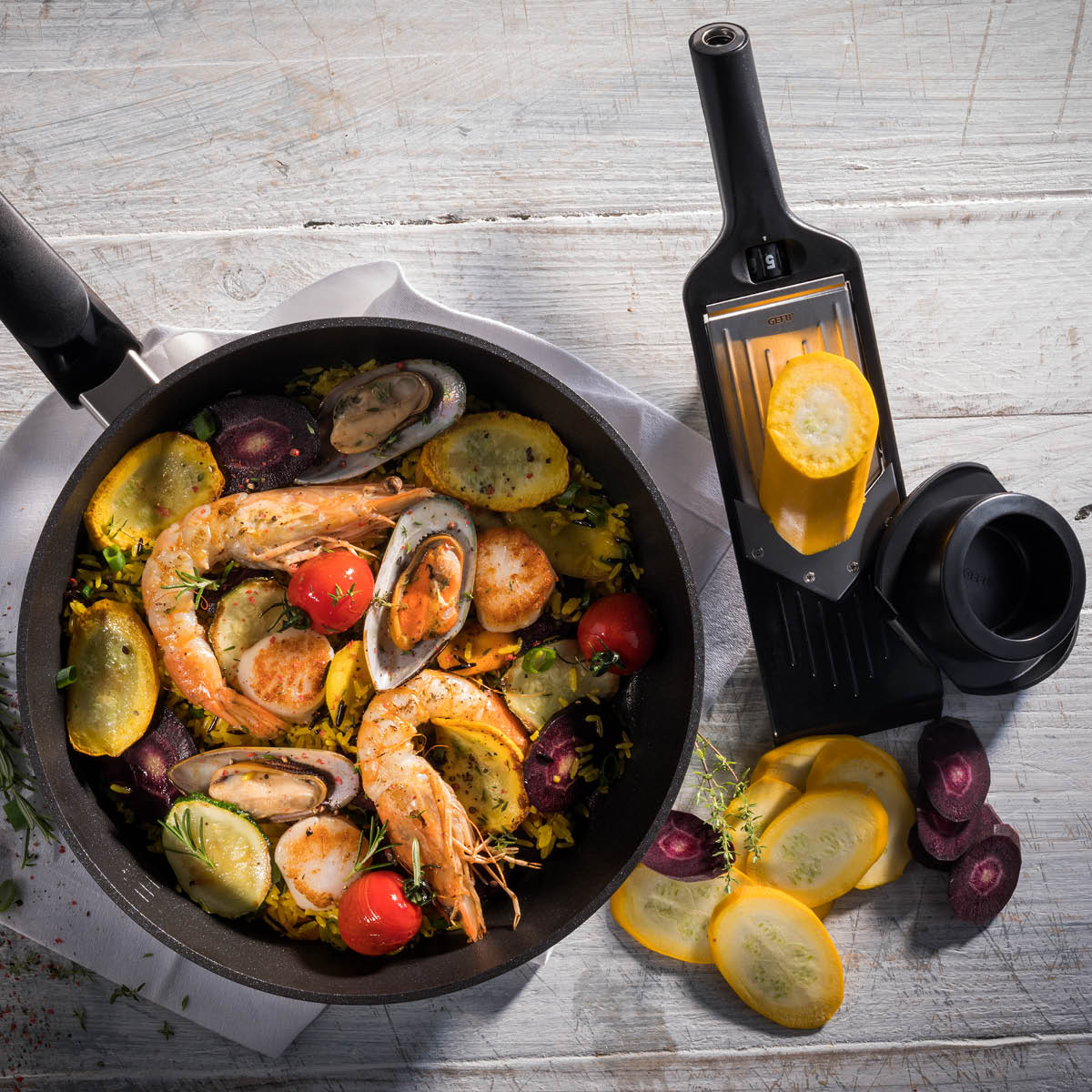 Frische Meeresfrüchte mit Gemüse in Pfanne neben Gemüsereibe auf rustikalem Tisch Pfanne mit Garnelen, Muscheln und Gemüse neben einer Reibe mit geschnittenem Sommergemüse auf Holztisch