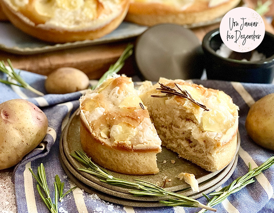 Vegane Kartoffel-Focaccia-Torten mit „Feta“ und Rosmarin Hausgemachtes Rosmarin-Kartoffelbrot mit einer Scheibe abgeschnitten auf einem Holzbrett, umgeben von frischem Rosmarin und Kartoffeln, im Hintergrund Salzschale auf gestreiftem Tuch