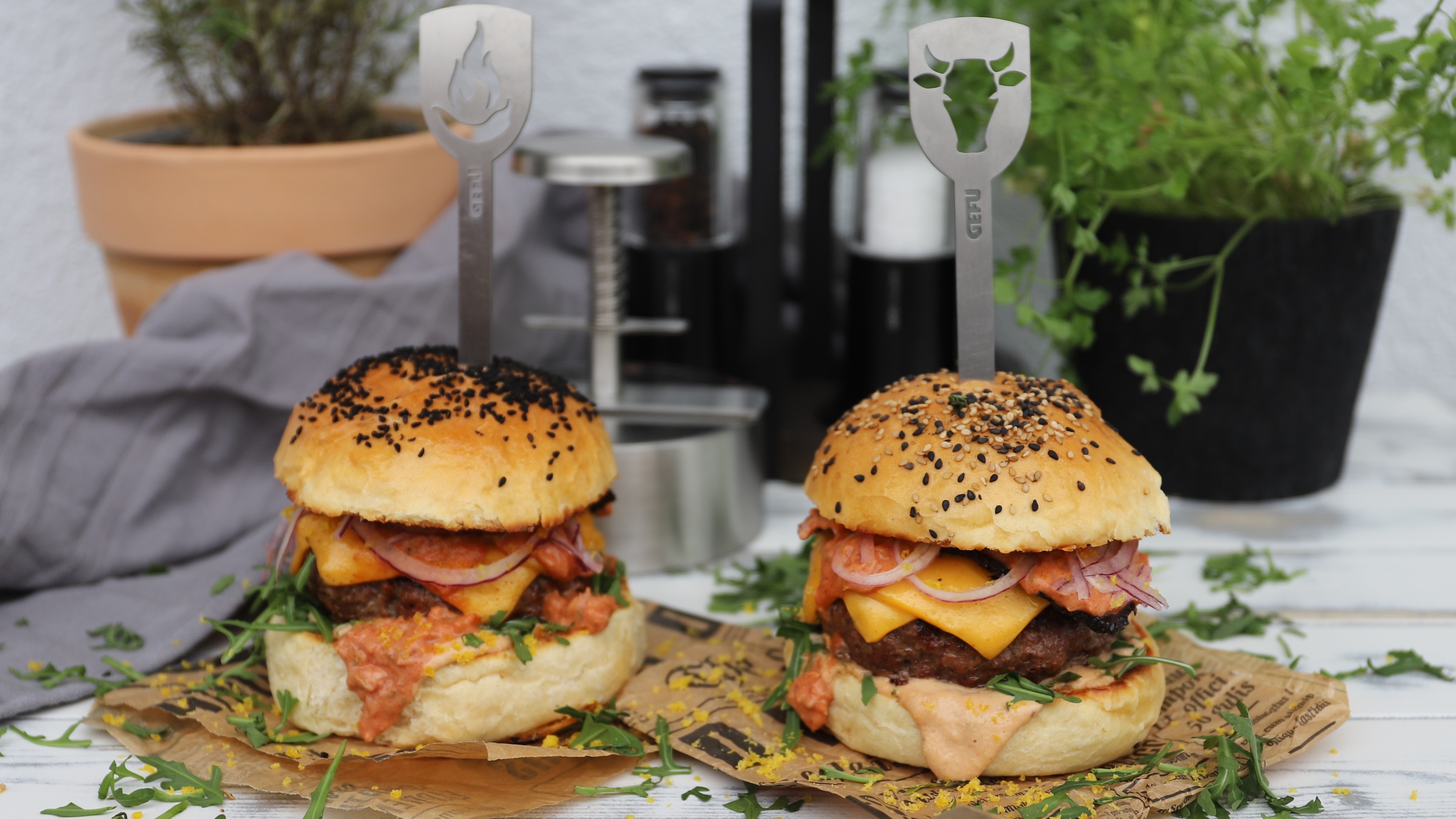 Hot-Chili Wagyu Burger mit Pastrami  Zwei saftige Cheeseburger mit Sesambrötchen, Salat, Käse, Speck und dekorativen Spießen serviert im Freien auf einem Holztisch mit Pflanzen im Hintergrund